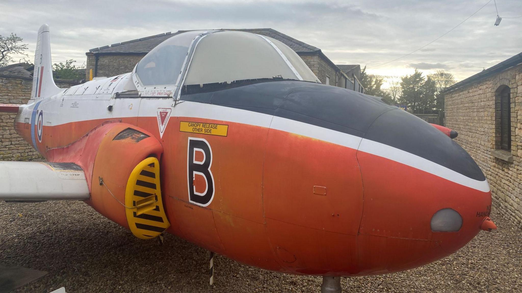 An old, weathered jet aircraft standing in a gravel yard outside yellow-brick buildings. The plane is painted orange on the bottom half and white on the top half. The top half of its nose is painted black. A letter large "B" is stencilled on to the side. The sky is grey.