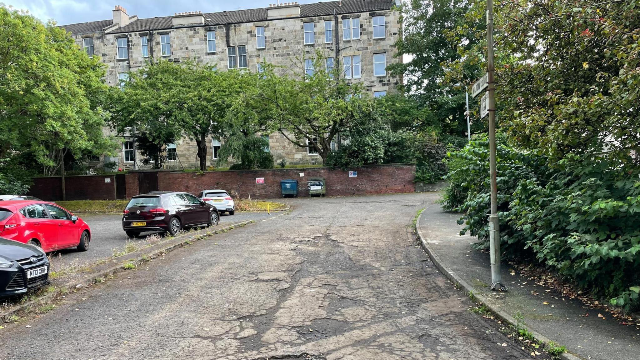 Potholed road with cars parked next to it. There are flats, trees and a street sign and two bins against a wall in the distance