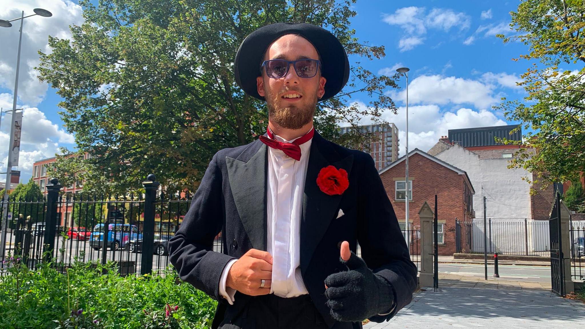 A man dressed in a hat and fingerless glove and bow tie shows the thumbs up sign