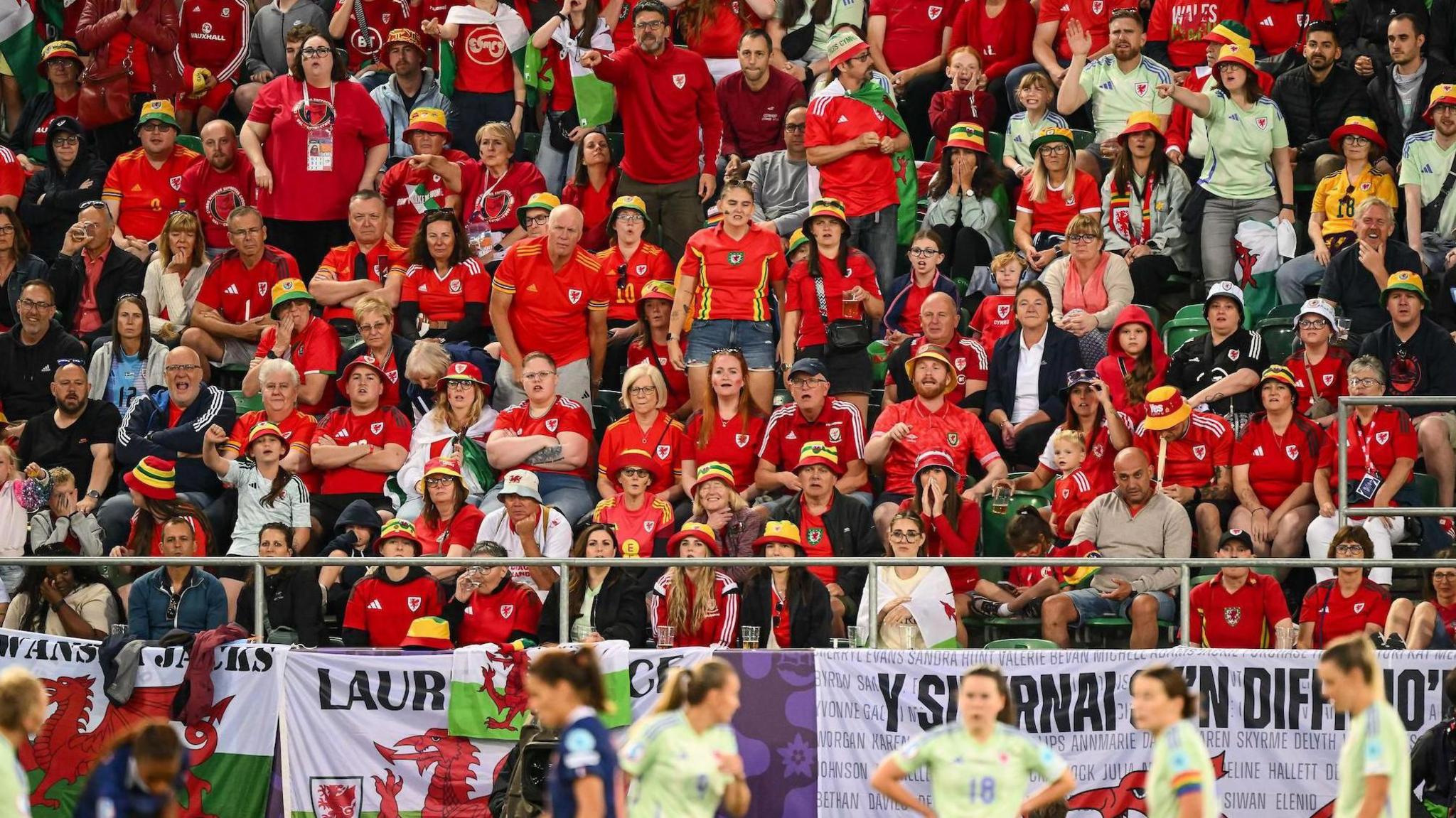 Wales fans in the stands watch Wales against France in St Gallen. Welsh national flags are draped over barriers with Wales players in lime green change shirts stand on the pitch.