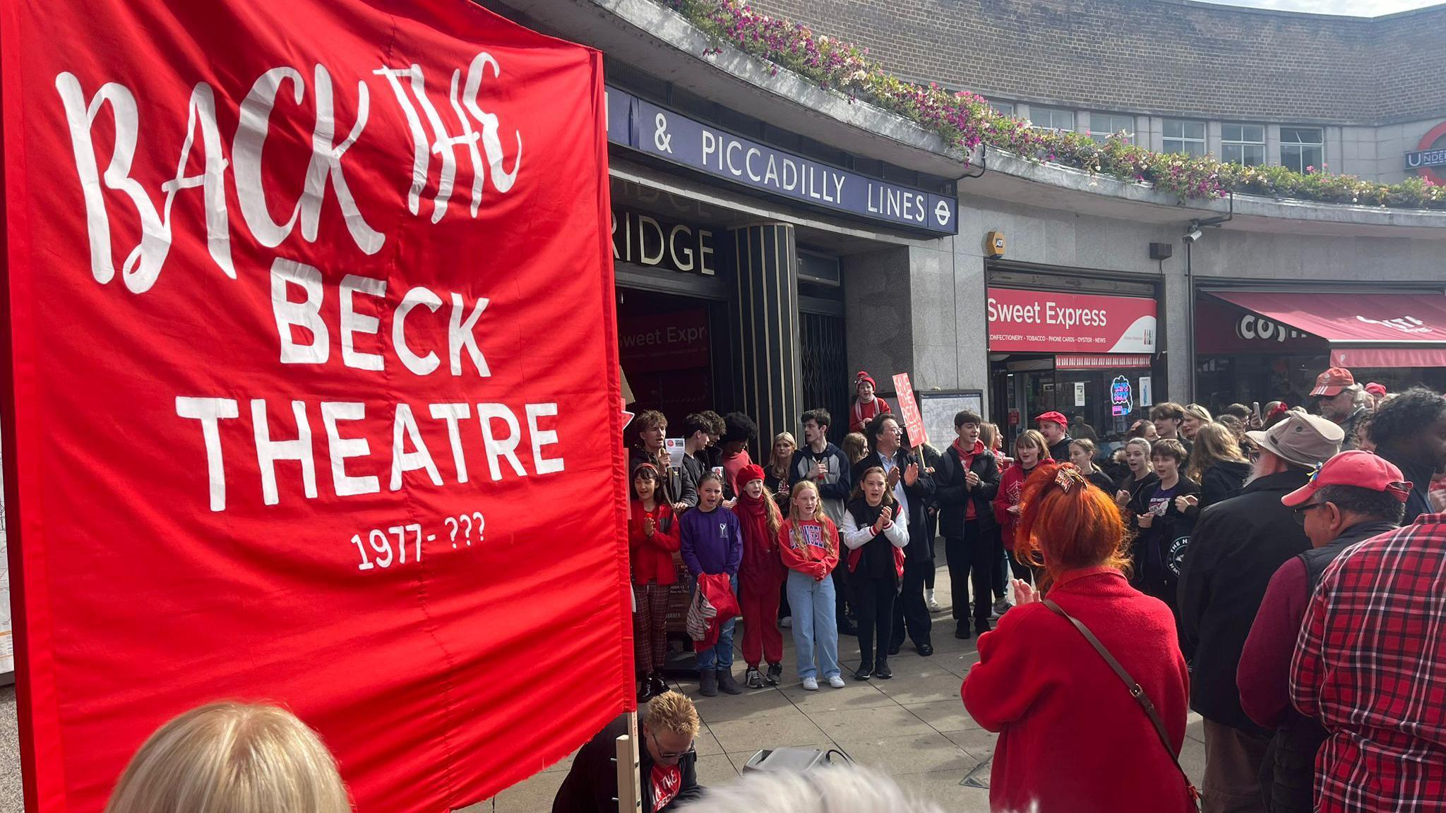 crowds of people holding 'back the beck theatre' banners outside the entrance to uxbridge tube station