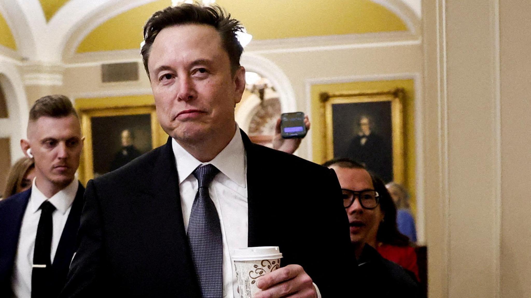 Elon Musk walking in Congress. The billionaire is holding a coffee cup as reporters follow behind him. 