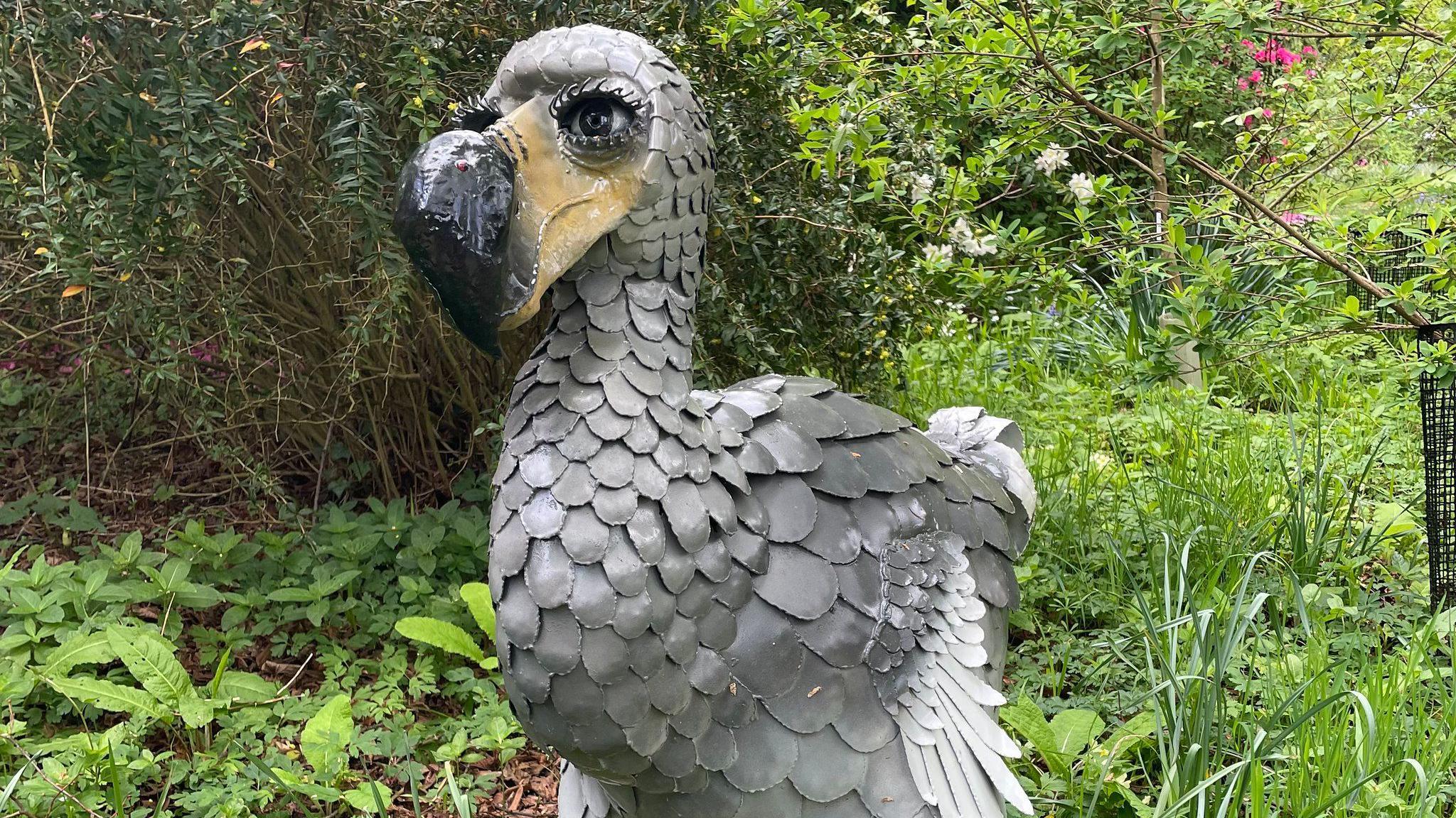 A statue of a grey bird with a yellow beak, surrounded by greenery. The bird has large, human-like eyes with prominent eyelashes.
