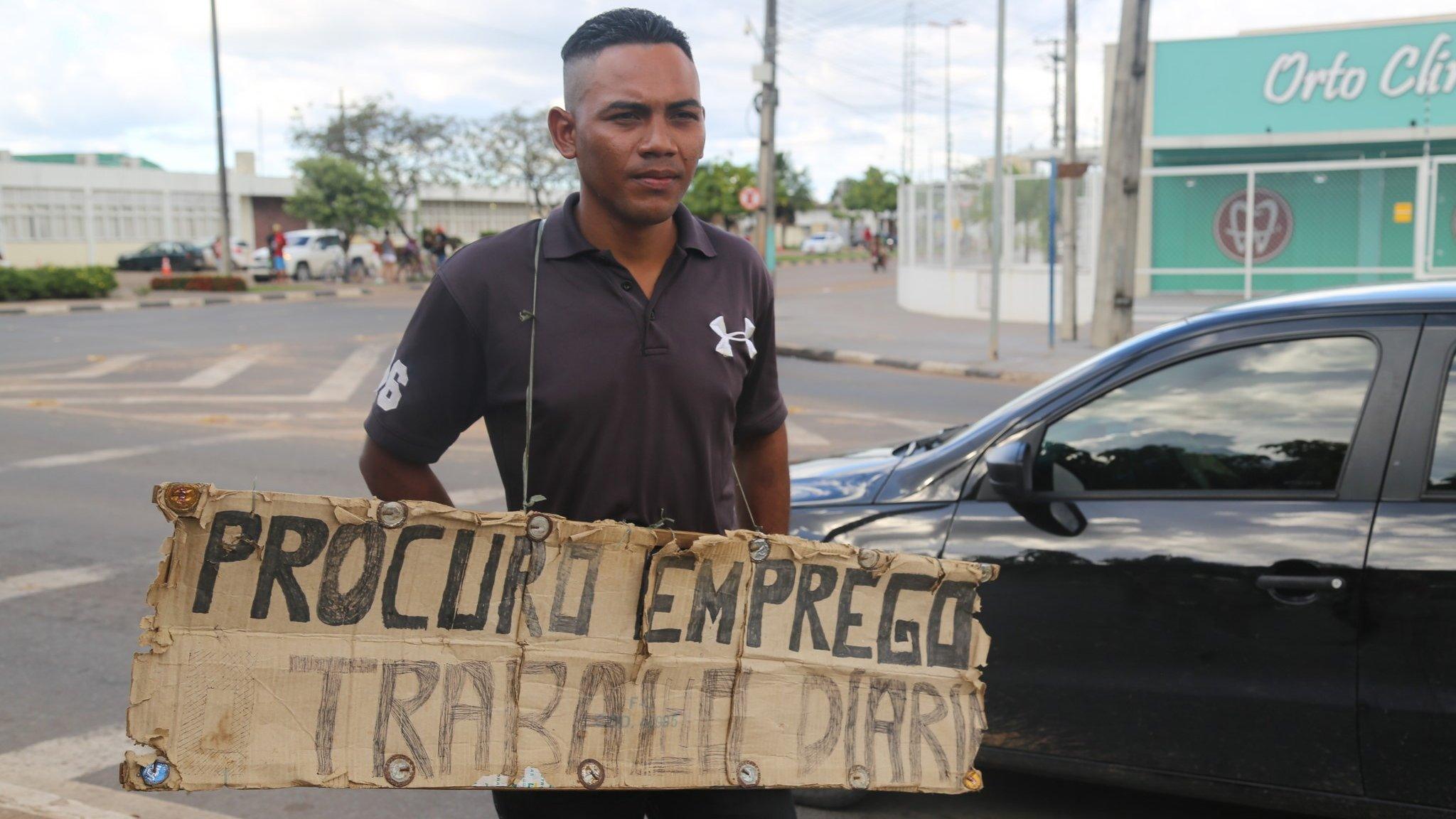 A man carries a sign reading "Looking for work" in Portuguese around his neck in Boa Vista