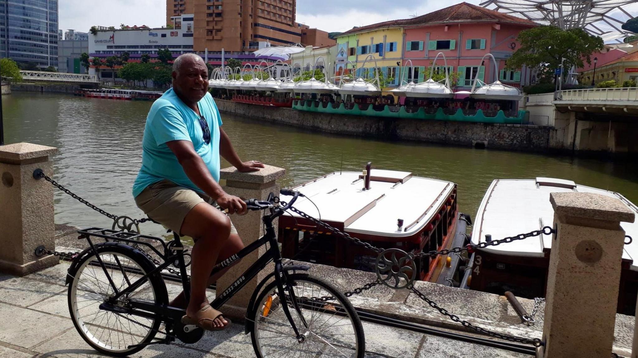 Julian Delaney on a bicyle by a river in a city in Singapore with colourful buildings in the background. He is wearing a blue T-shirt with tan coloured shorts and brown sandals. His bike is all black 