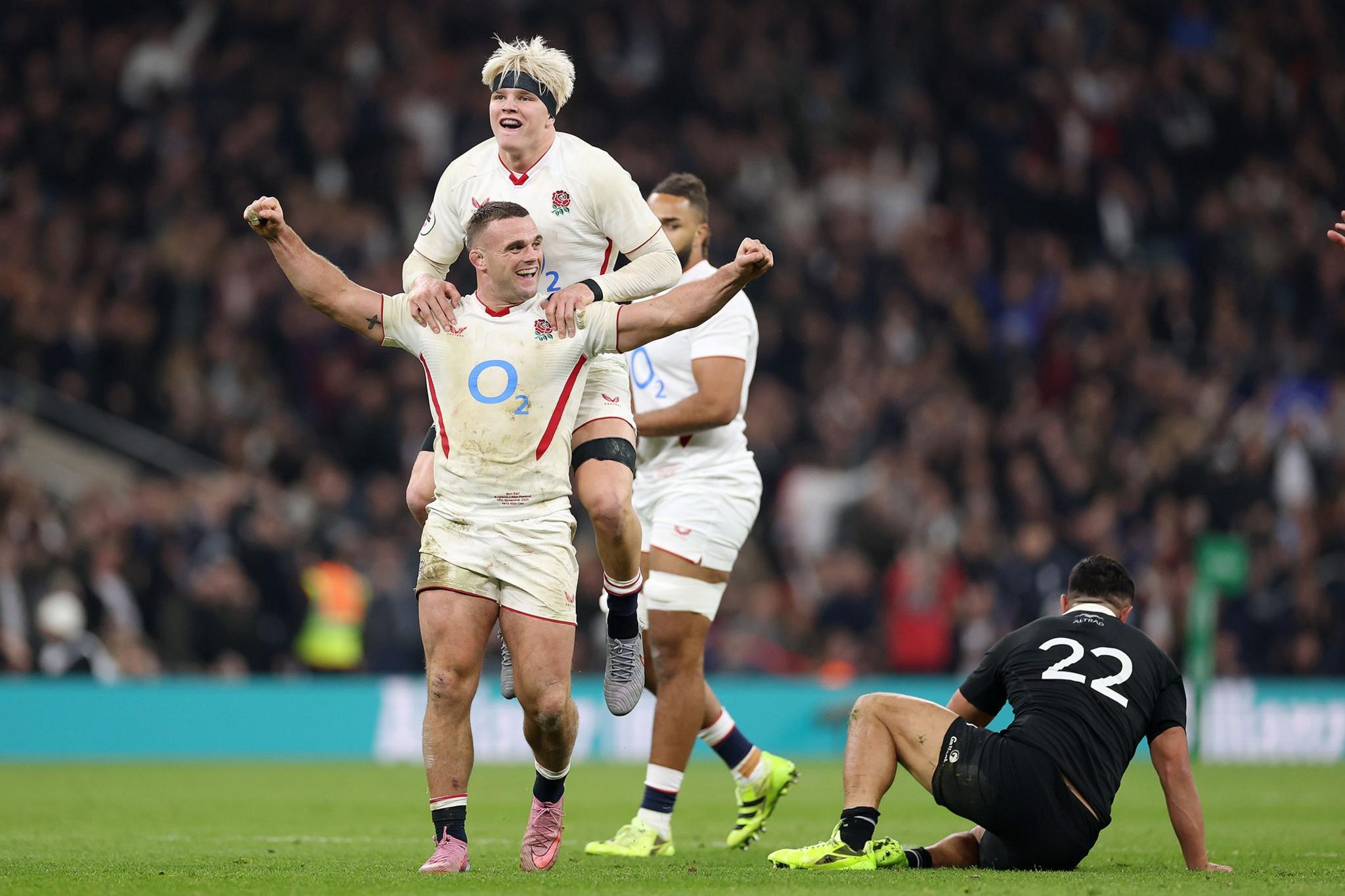 Rugby players in white England kits celebrating on the field, one lifting another while raising arms in triumph, with an opposing player in black sitting on the ground in the background.
