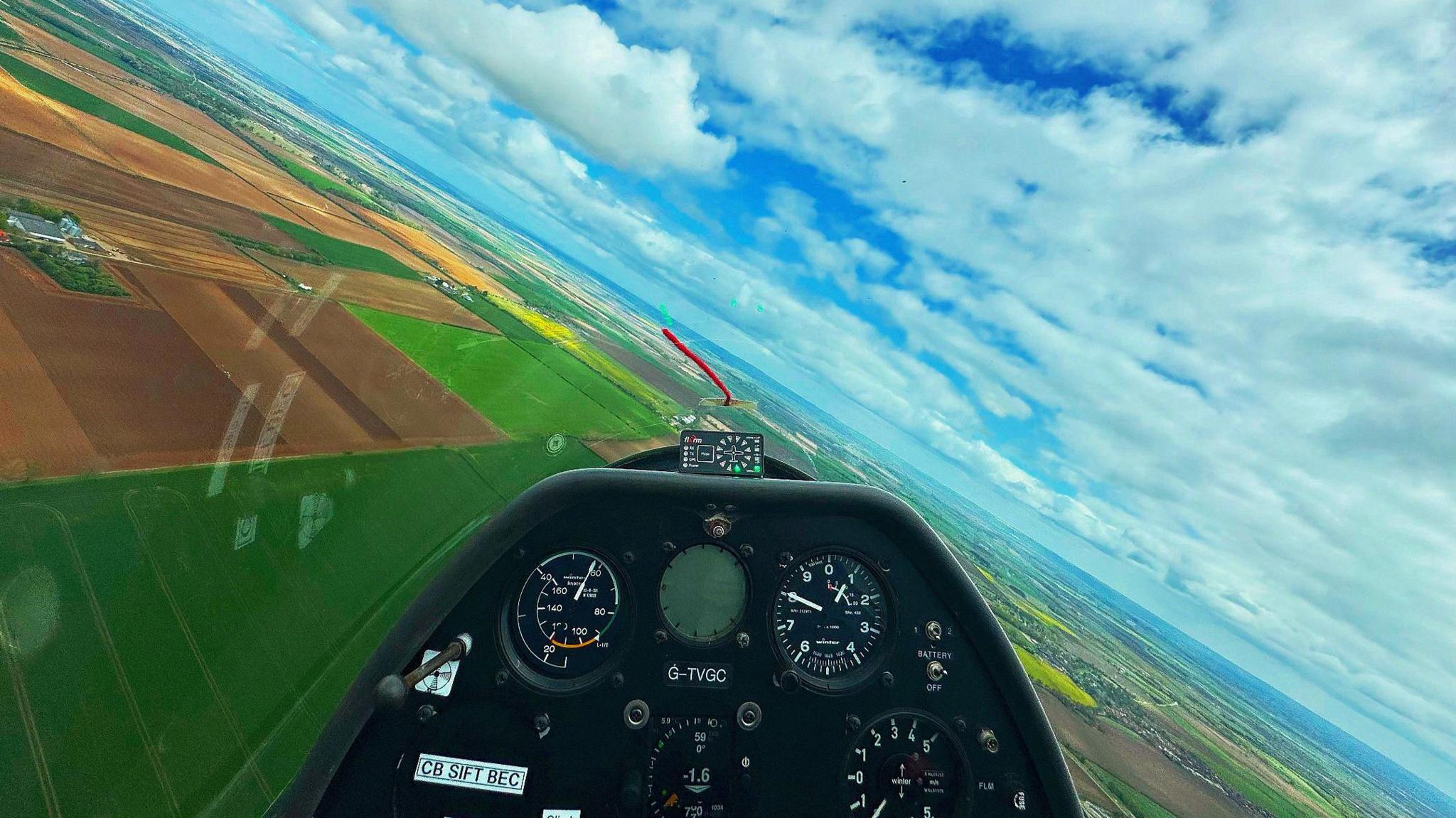An aerial view of inside a glider, dials and buttons are in front. The glider is above many fields which can be seen far and wide, from lush green crops to ploughed fields. In the distance some villages can be seen. Above is a blue sky with clouds.