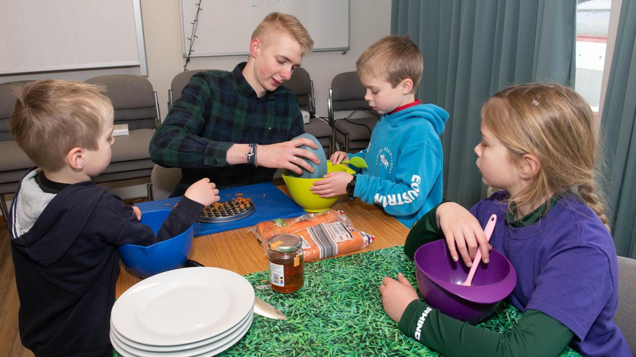 Three children with brightly coloured plastic food bowls learn how to bake, taught by a young person.
