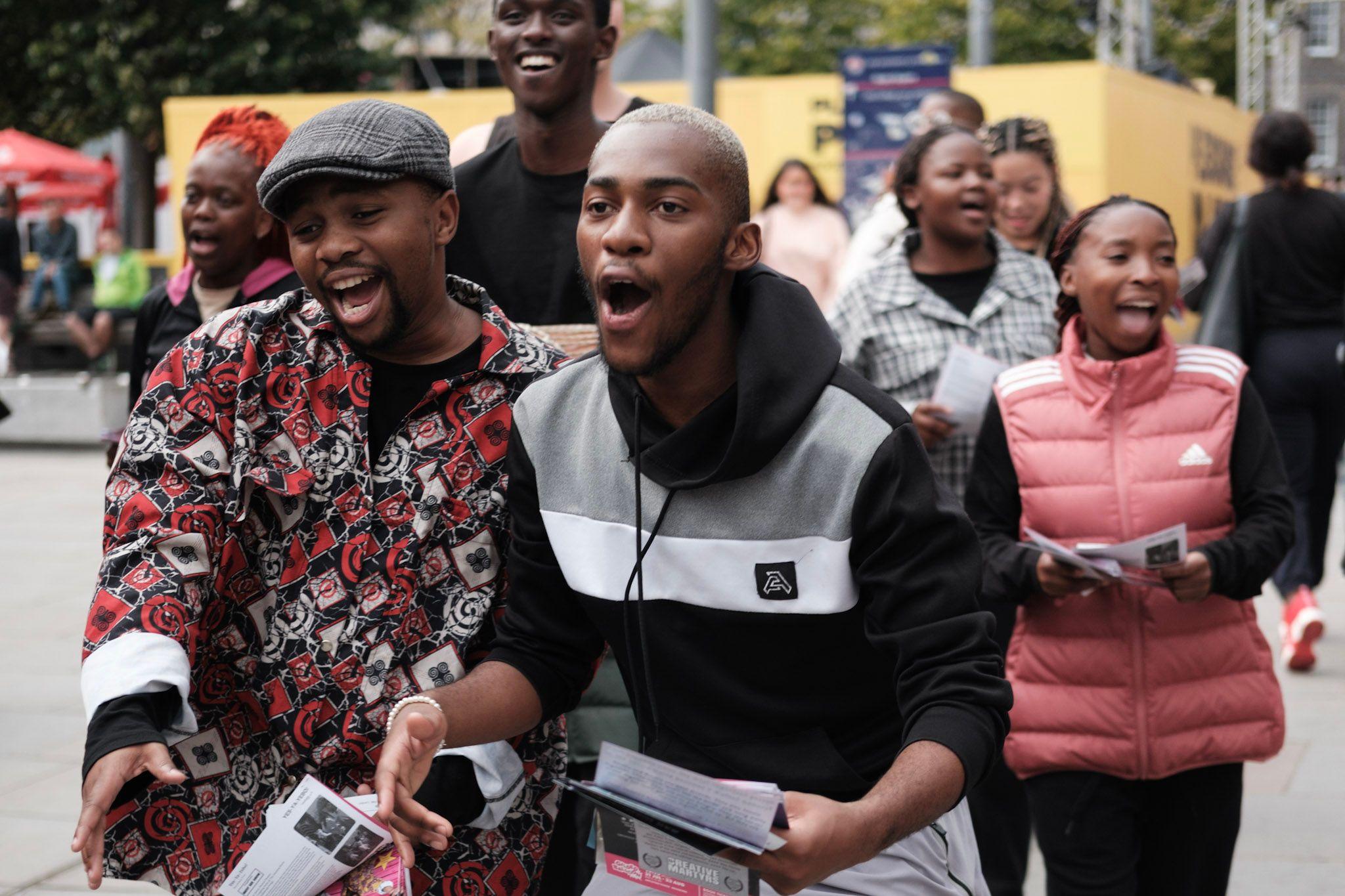 A group of performers sing as they hand out leaflets. They are young and wear trendy clothes.