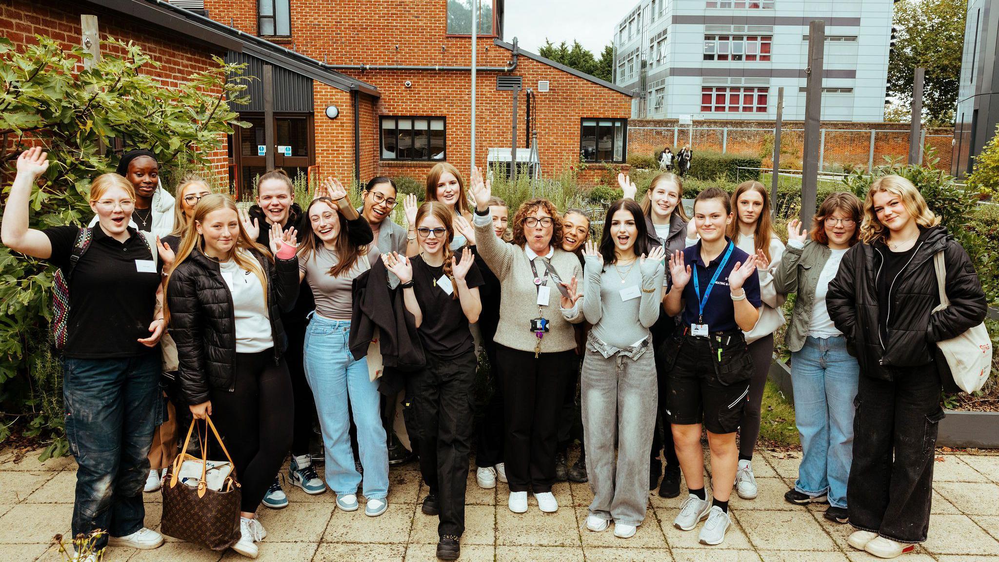 The seventeen female students from West Suffolk College are pictured. They are in a close group facing the camera. They all have their hands up celebrating with smiles on their face. Buildings from the college can be seen behind them.