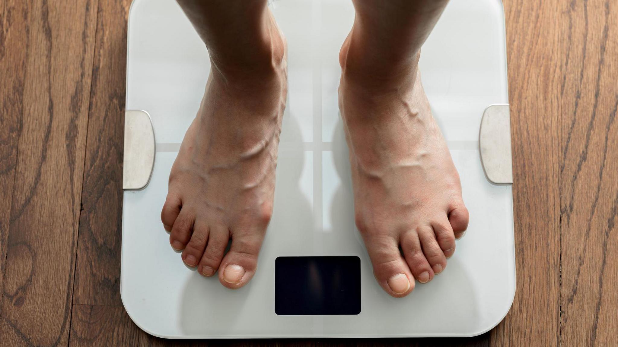 The feet of a person standing on white bathroom scales with a black digital screen in between the feet. 