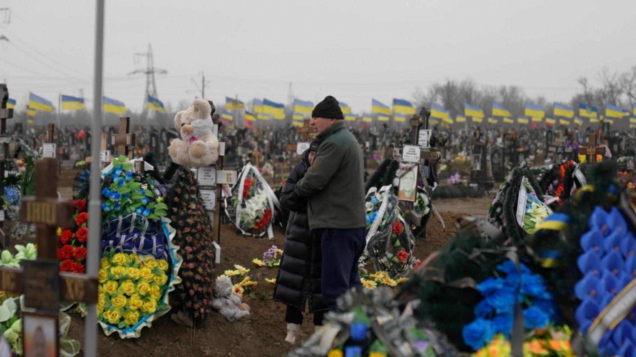 Yuliia Tarasevych and Serhiy Lushchay embrace in a cemetery filled with Ukrainian flags.  