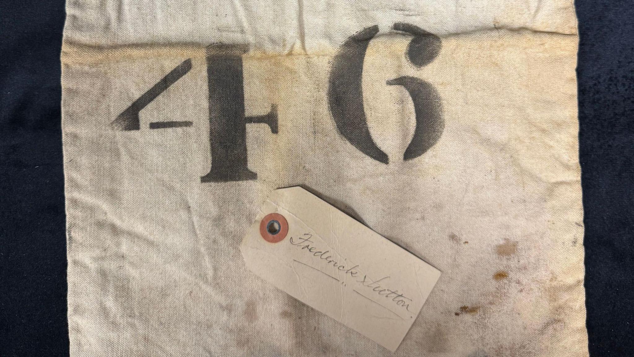 An old, white canvas bag with a large number 46 on it and a tag bearing the name Frederick Sutton in cursive handwriting.