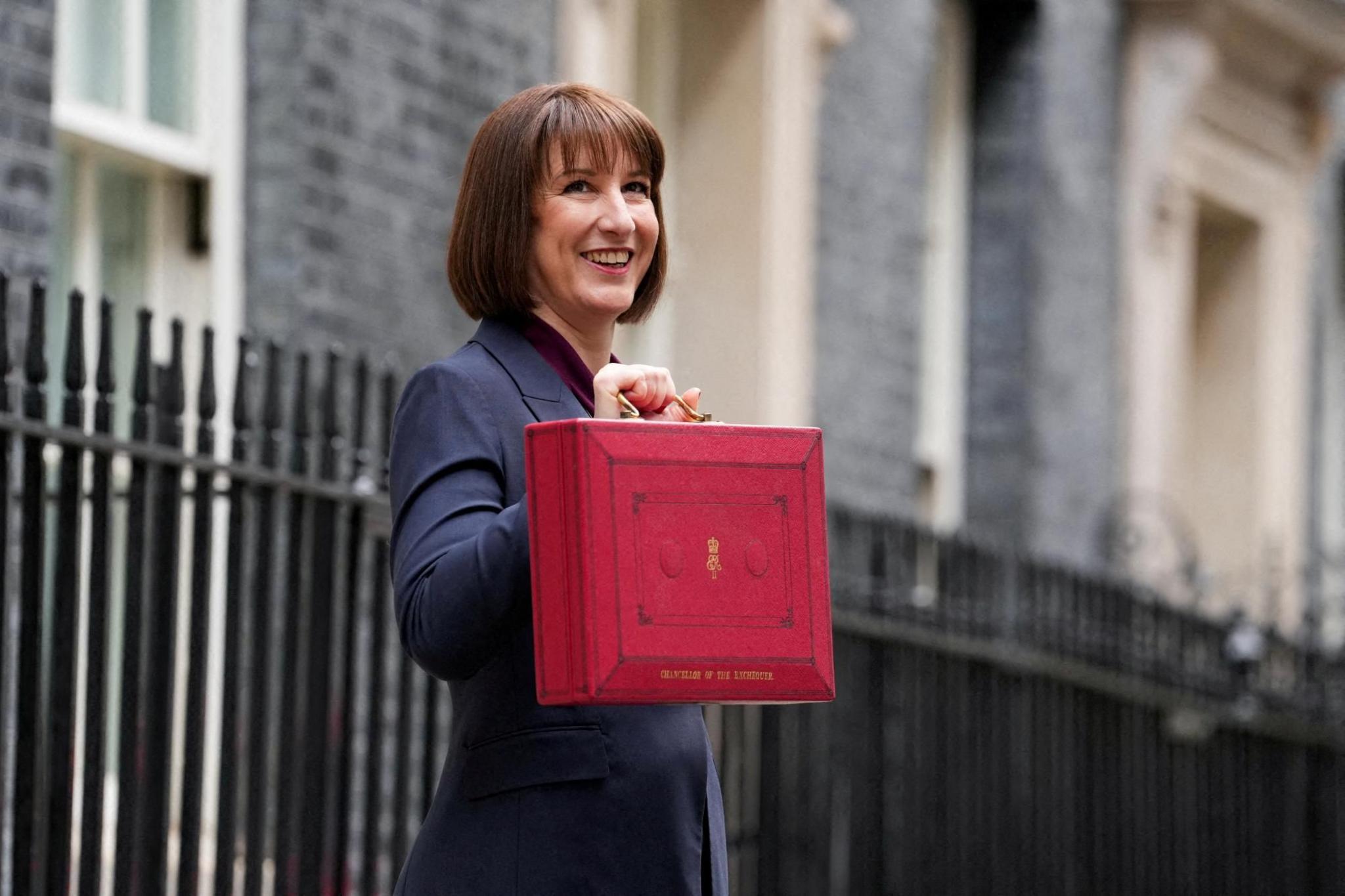 Rachel Reeves standing in front of black railings holding her red Budget box. She is smiling and wearing a dark blue suit jacket.