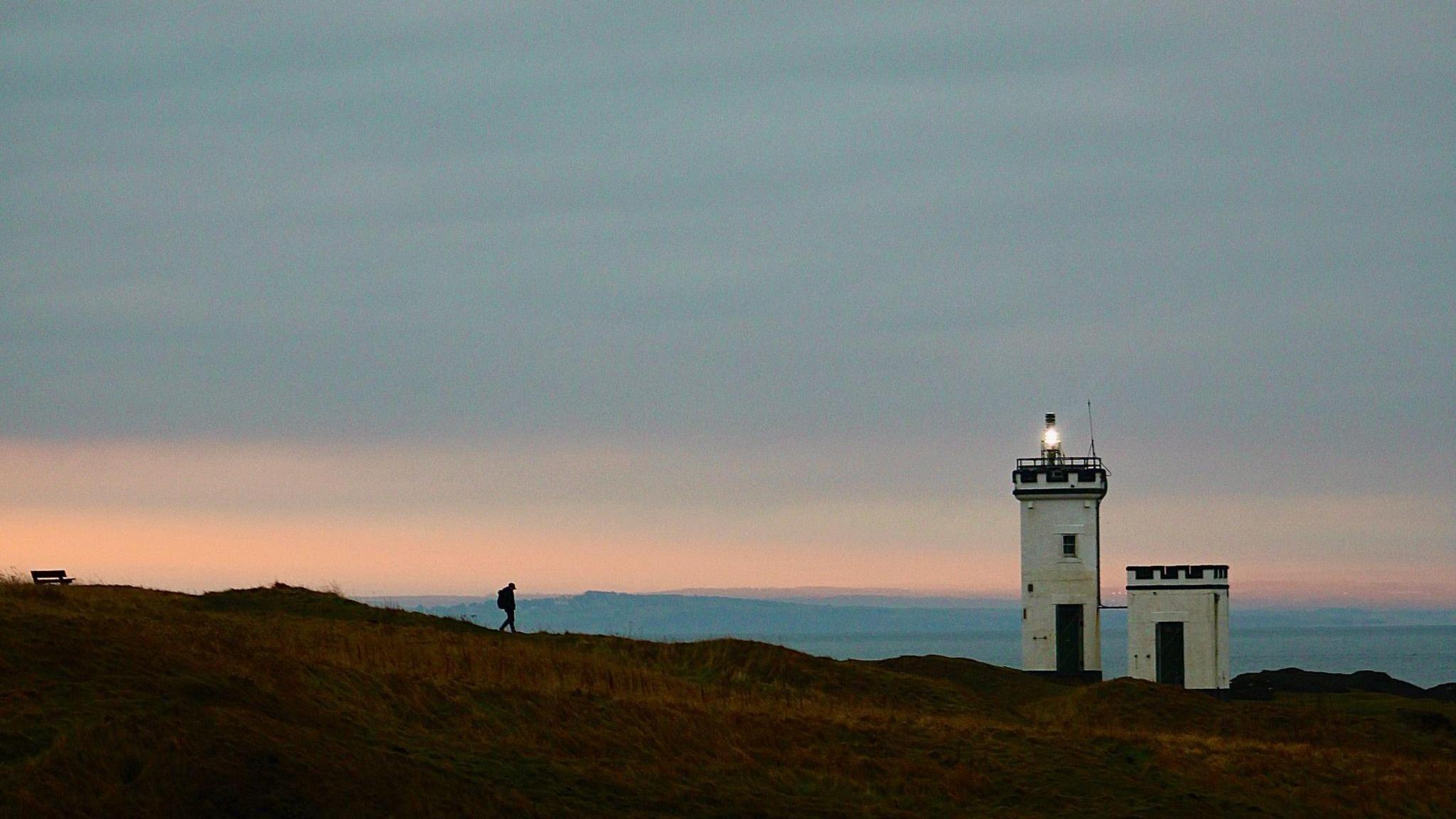 The silhouette of a person walking towards a lighthouse in front of a blue and pink sky