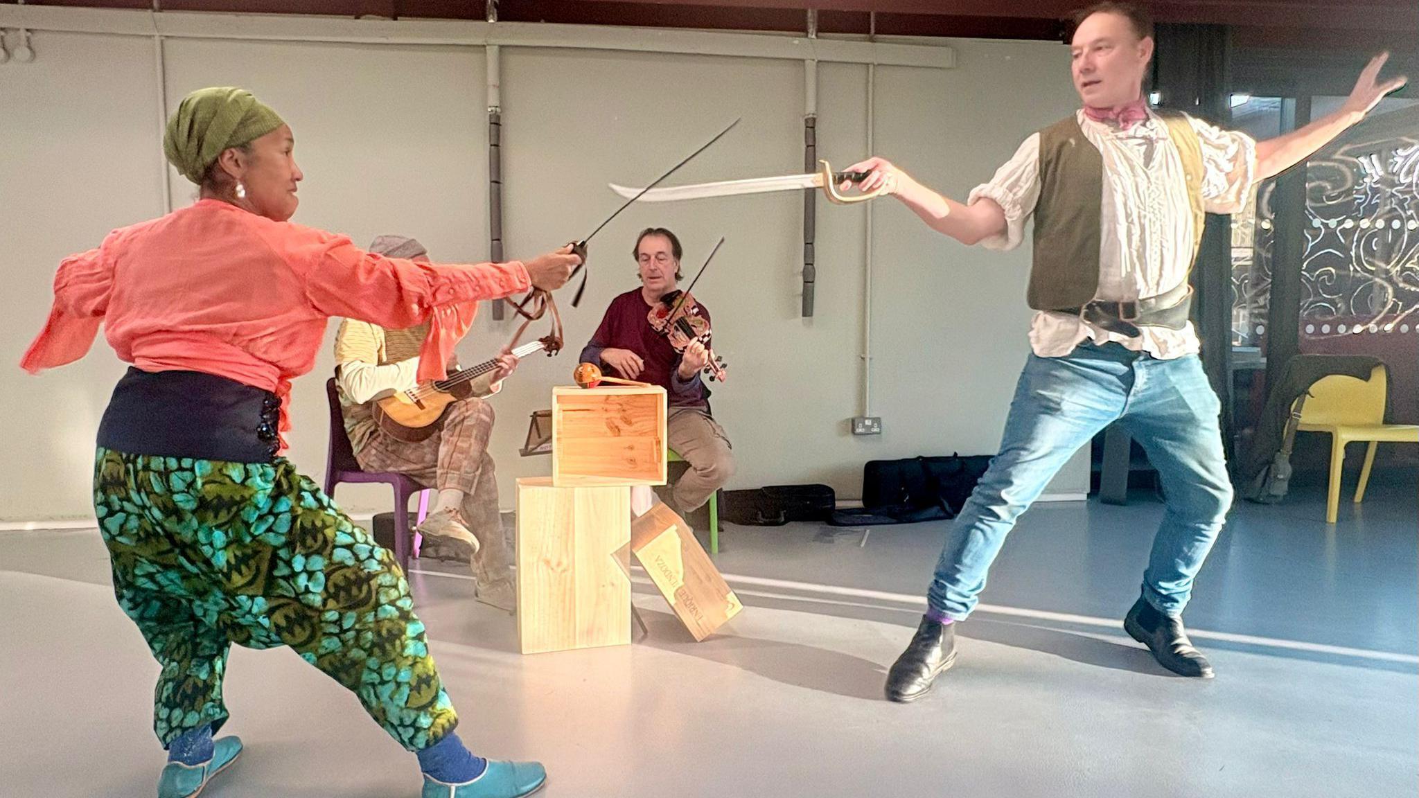 Mary-Ann Roberts a Richard Parry mewn ymarferion yn dal cleddyfau. Mae dau ddyn yn chwarae offerynnau yn eistedd yn y cefndir.