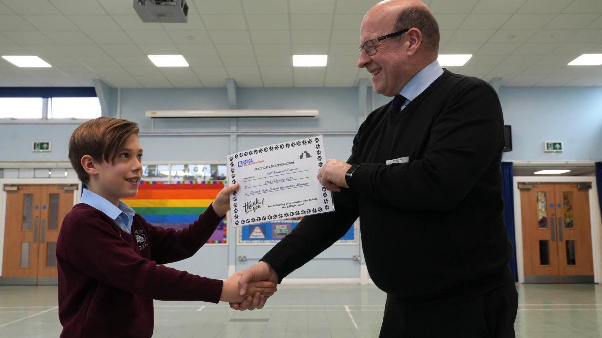 A young boy in school uniform receives an award and shakes the hand of a man from the RSPCA. They appear to be standing in a sports hall.