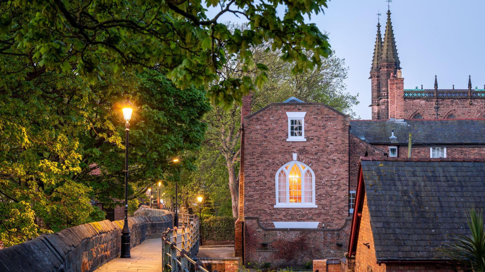 Streetlamps are glowing along a lined section of the walls. Bricked buildings can be seen with the Cathedral pictured at the top of the image. Green leafy trees overhang on to the walkway from above.