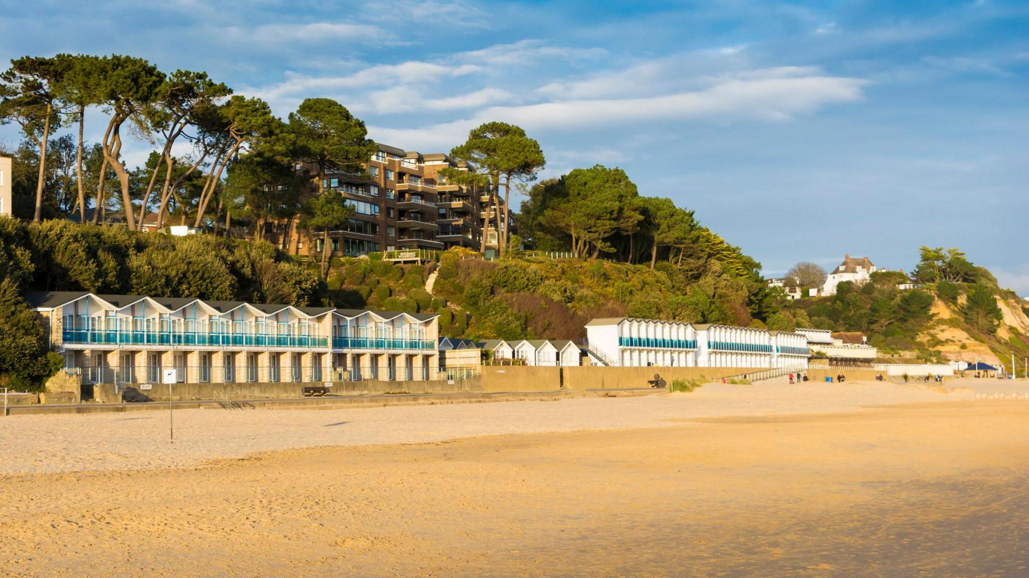 A row of beach huts on the promenade next to a sandy beach. Some of the rows are two-tier. They have blue glass balconies. Behind the beach huts is green vegetation and trees, with a building which looks to be a block of flats or a hotel on the clifftop. The sun is giving a slight orange glow to everything