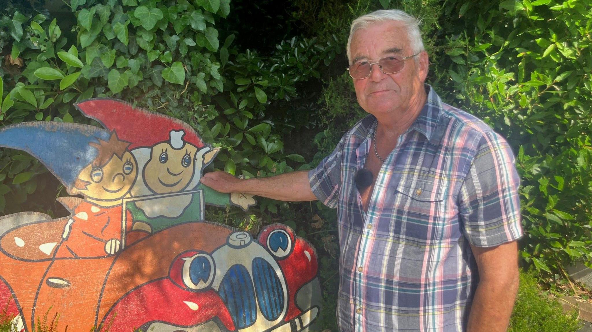 A man stands next to a plywood sign painted with the children's characters Noddy and Big Ears. He wears a short sleeve shirt.