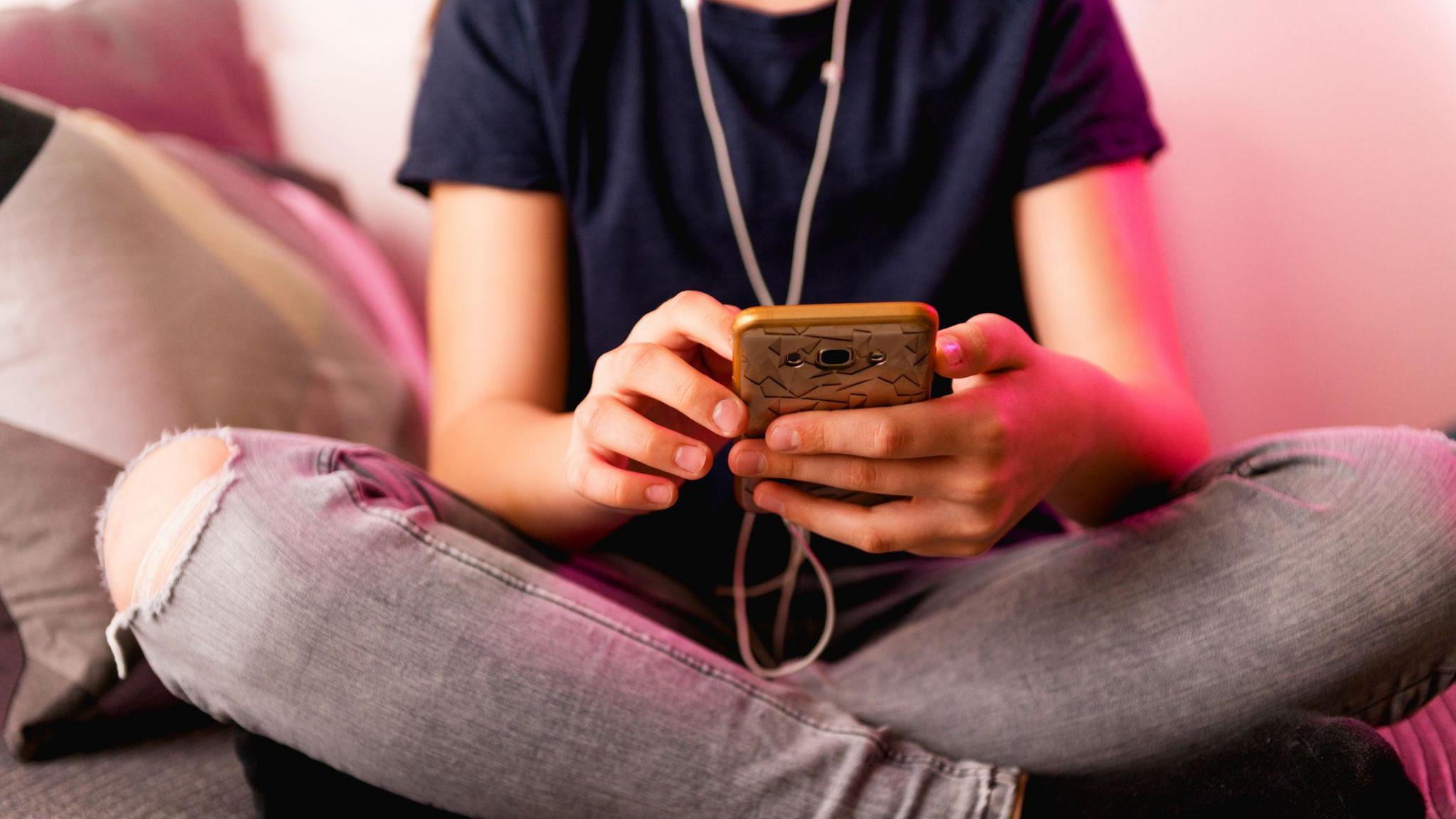 A teenage girl in a blue top and grey jeans sits cross-legged on an armchair, holding a mobile phone in her hands