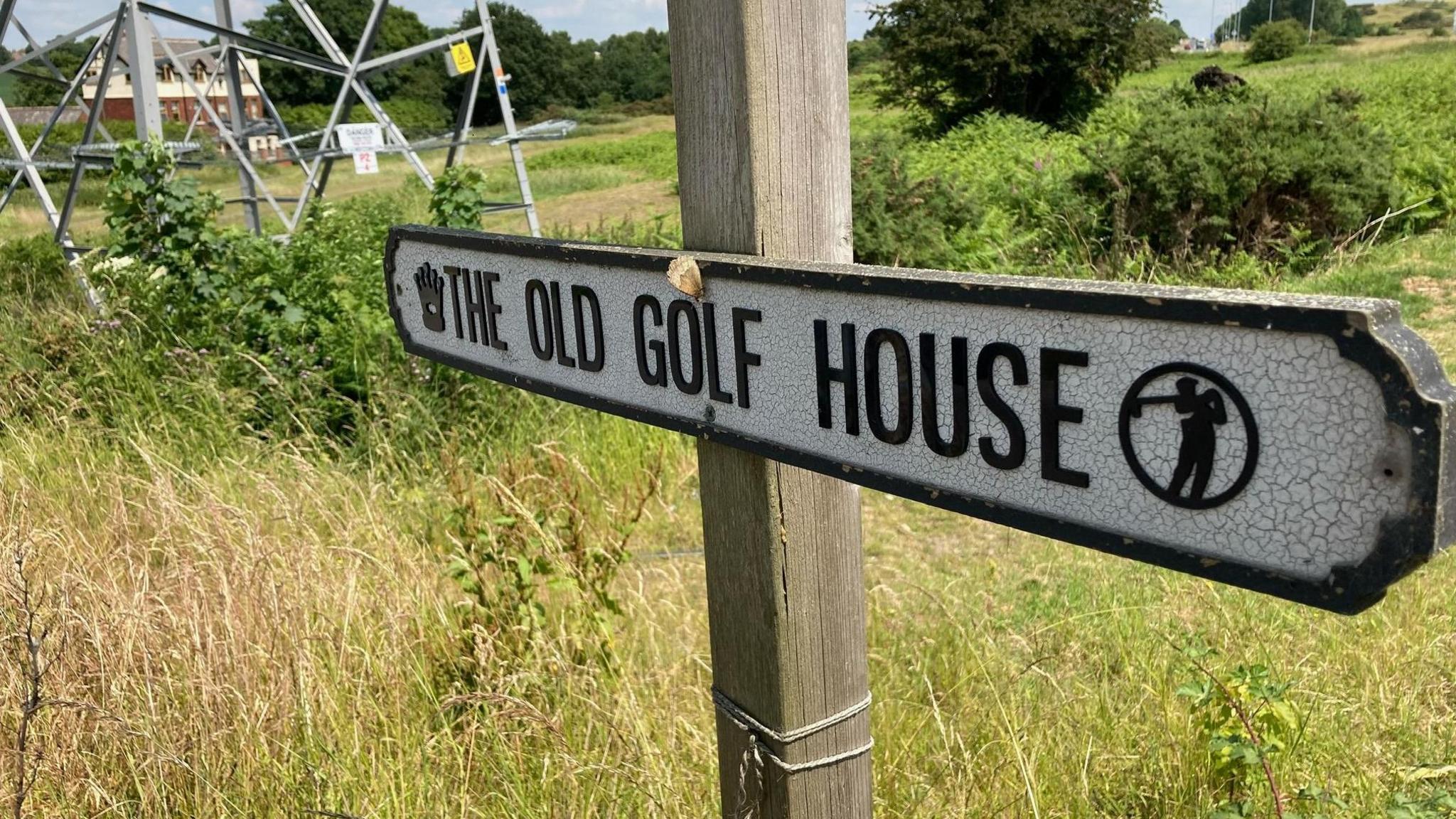 A sign bearing the words 'The Old Golf House' sits horizontally across a vertical wooden poll, against the backdrop of some long grass, hedgerows and the bottom of an electric pylon.