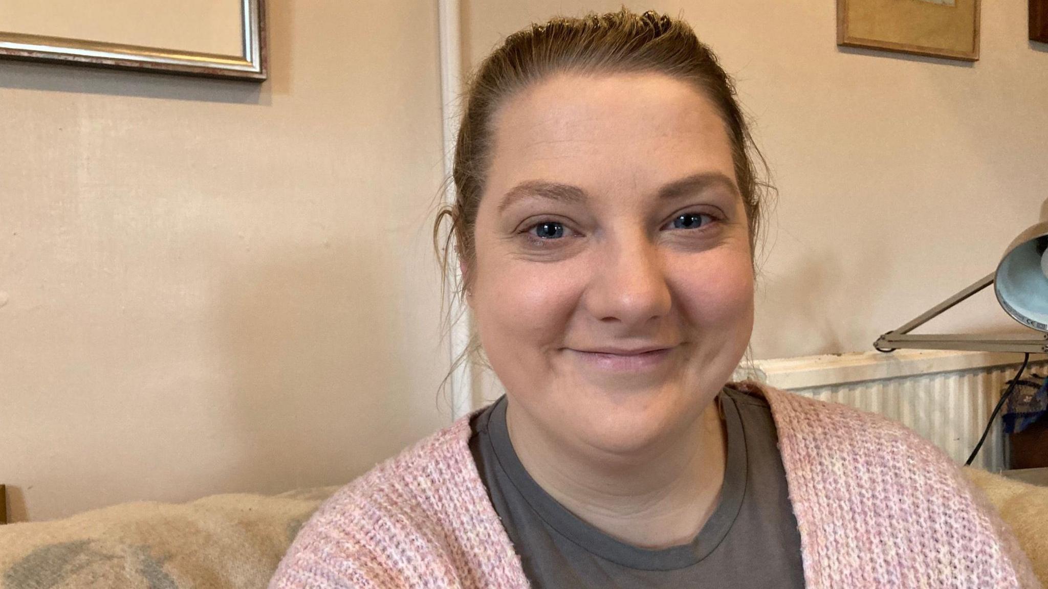 Florence Stokes looks at the camera while sitting on a sofa. She has dark blonde hair which is tied up. She is wearing a grey top and a light pink cardigan. She is looking directly at the camera and smiling. 