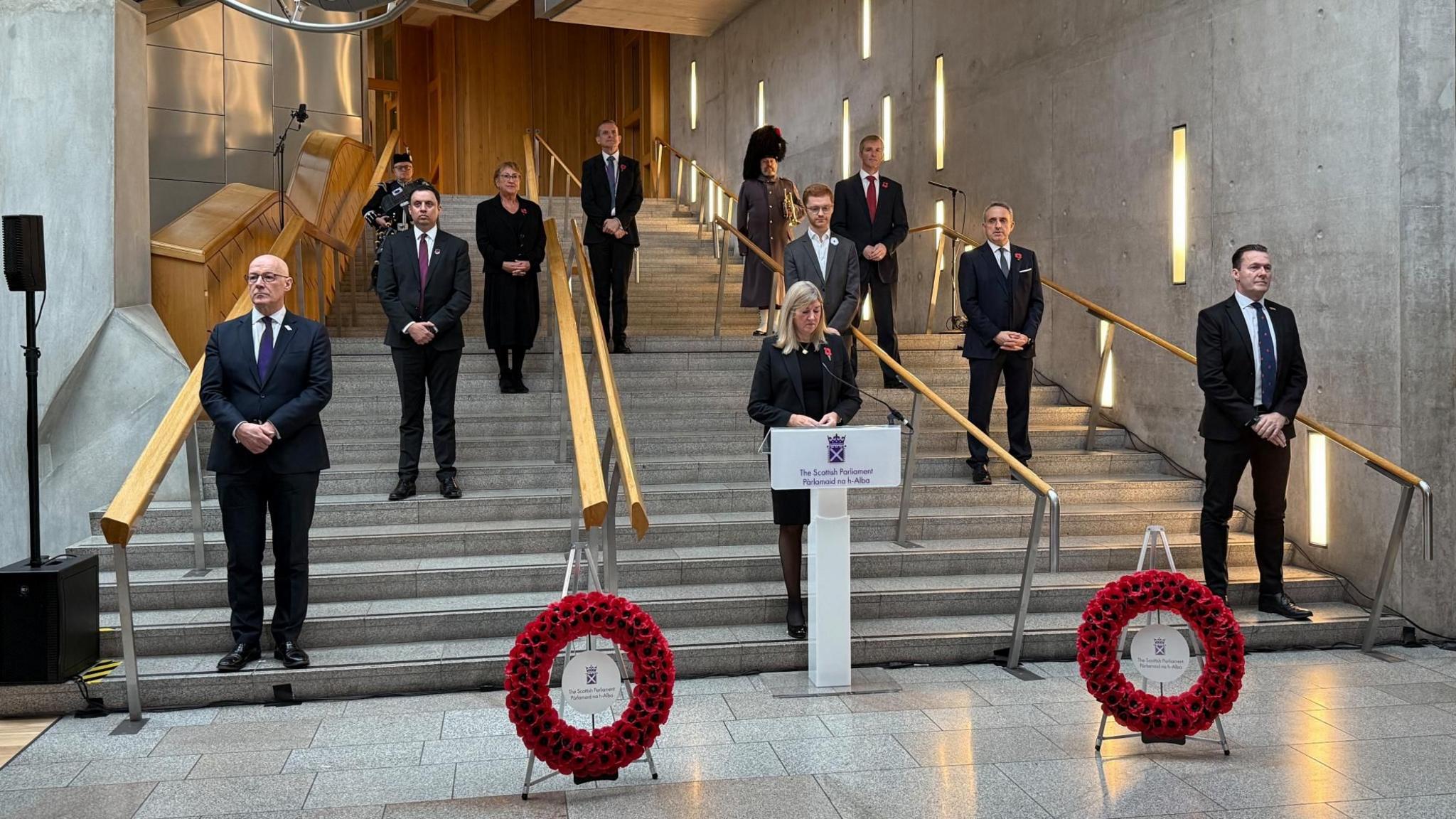 Members of Scottish Parliament dressed in formal attire stand on a staircase in Holyrood in silence paying tribute as a women stands at a podium. 