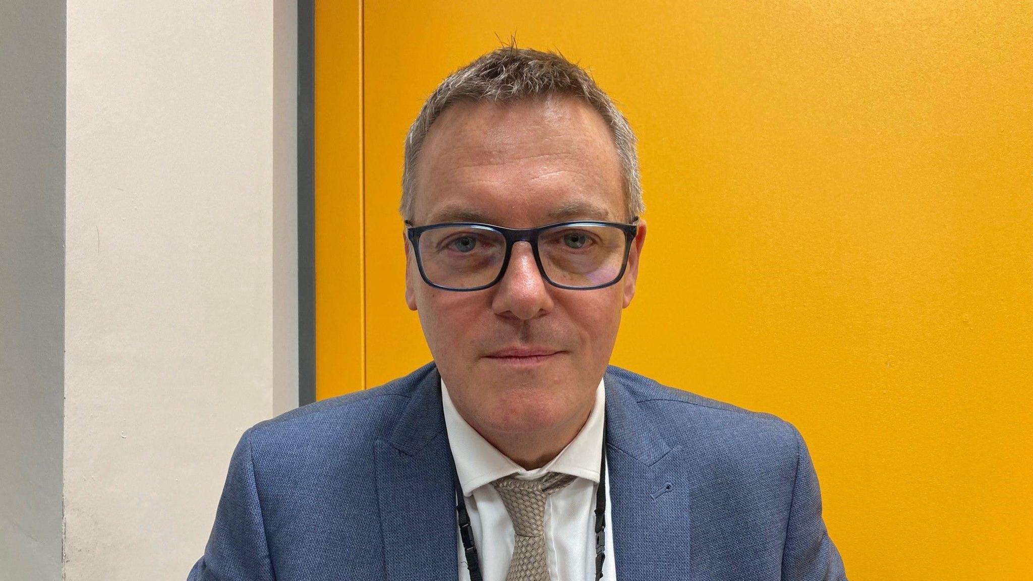 A man wearing glasses and a blue suit jacket and white shirt with a light brown tie in front of yellow background.