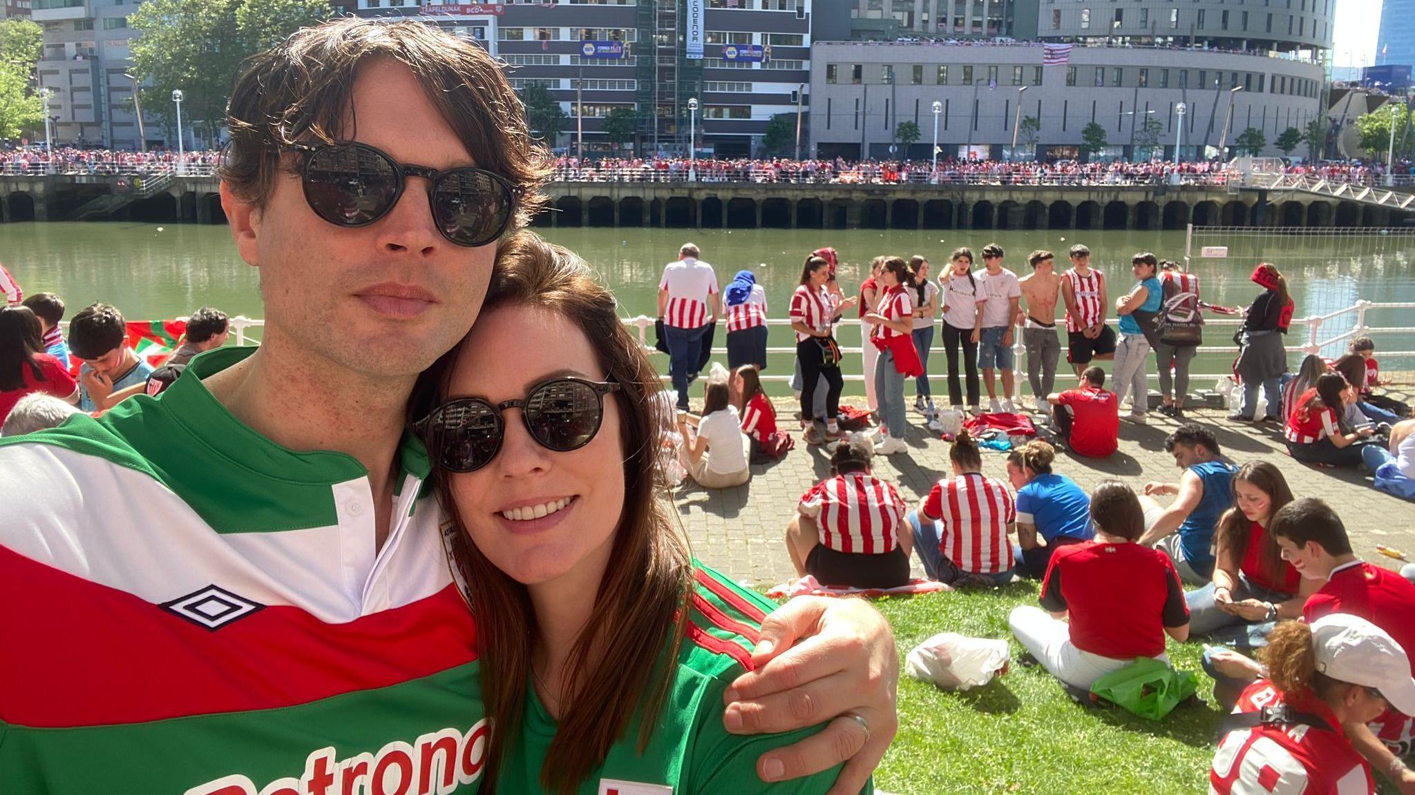 A man takes a selfies with a woman. He has dark brown hair and sunglasses on and a green, white and red striped football shirt, he has his arm around the woman who has a green shirt on with long dark hair and sunglasses. She smiles at the camera. Behind them there are people in red and white striped shirts sitting and standing by the river.