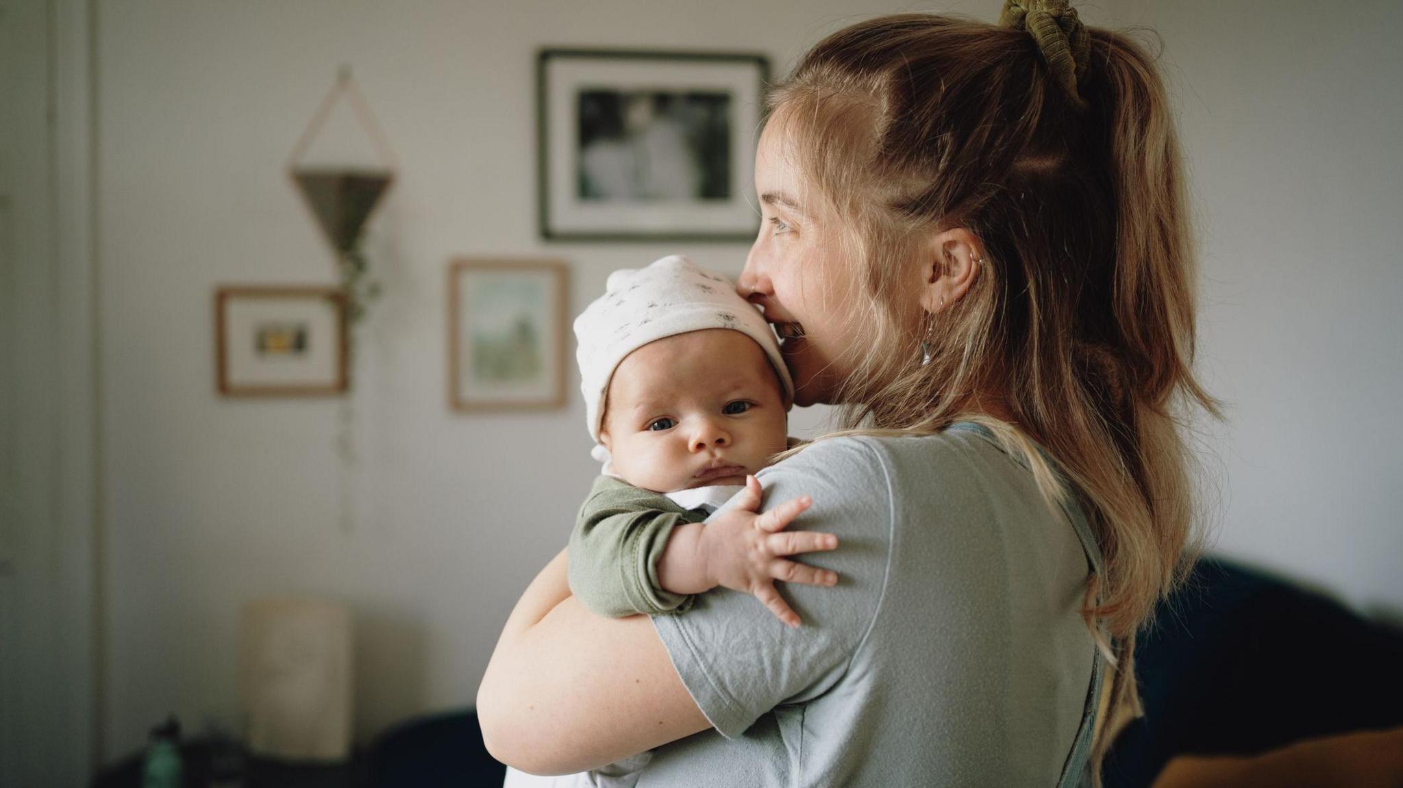 Mum with long blonde hair holding baby who is looking at camera with fingers outstretched. Picture posed by models.