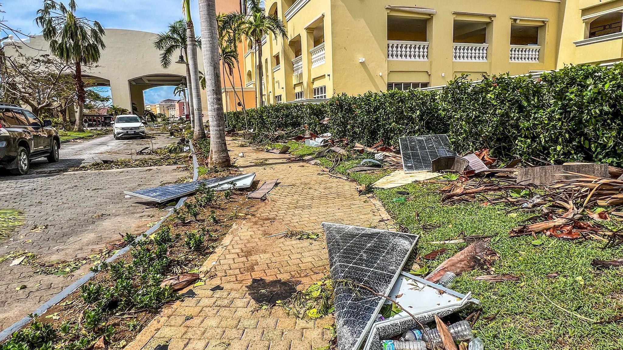A yellow building with palm trees outside is littered with broken solar panels and ripped up shrubs