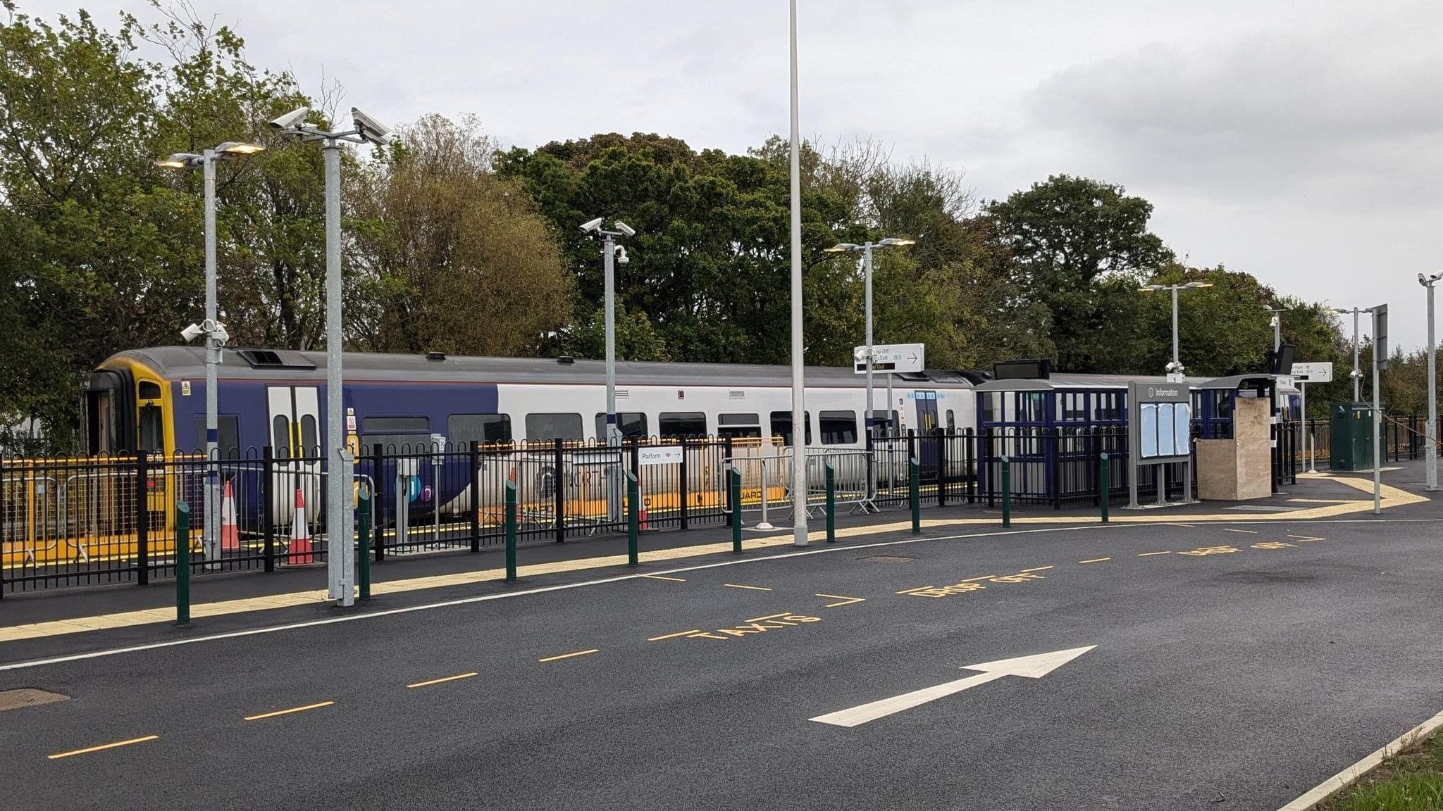 A two-carriage train stands at a new Ashington Station with black railings and a large carpark.