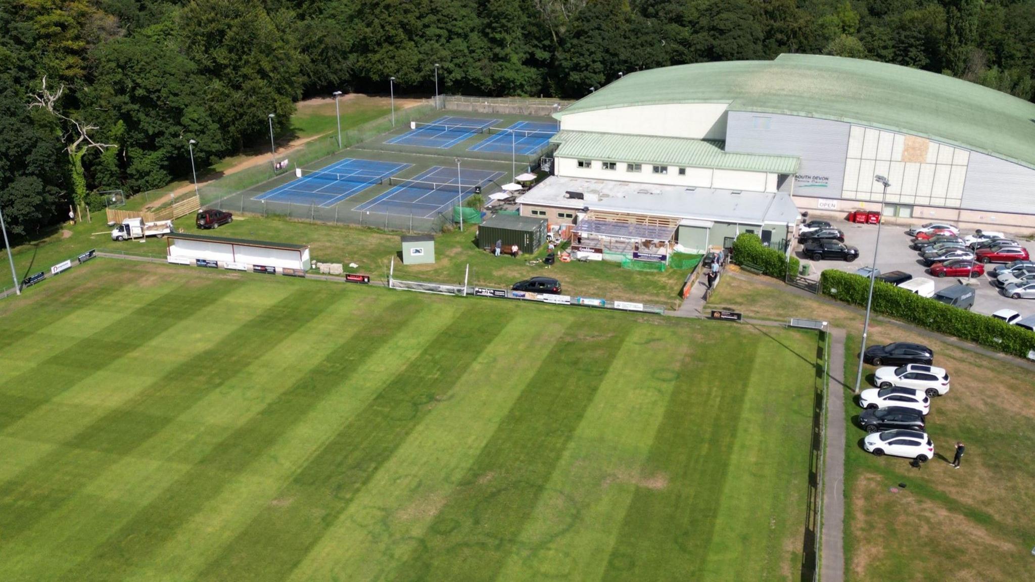 An aerial drone shot of the ground on a clear sunny day. It shows the clubhouse, part of the pitch, some tennis courts and a large building in the background. There is lots of trees and cars parked in a car park also in the background.