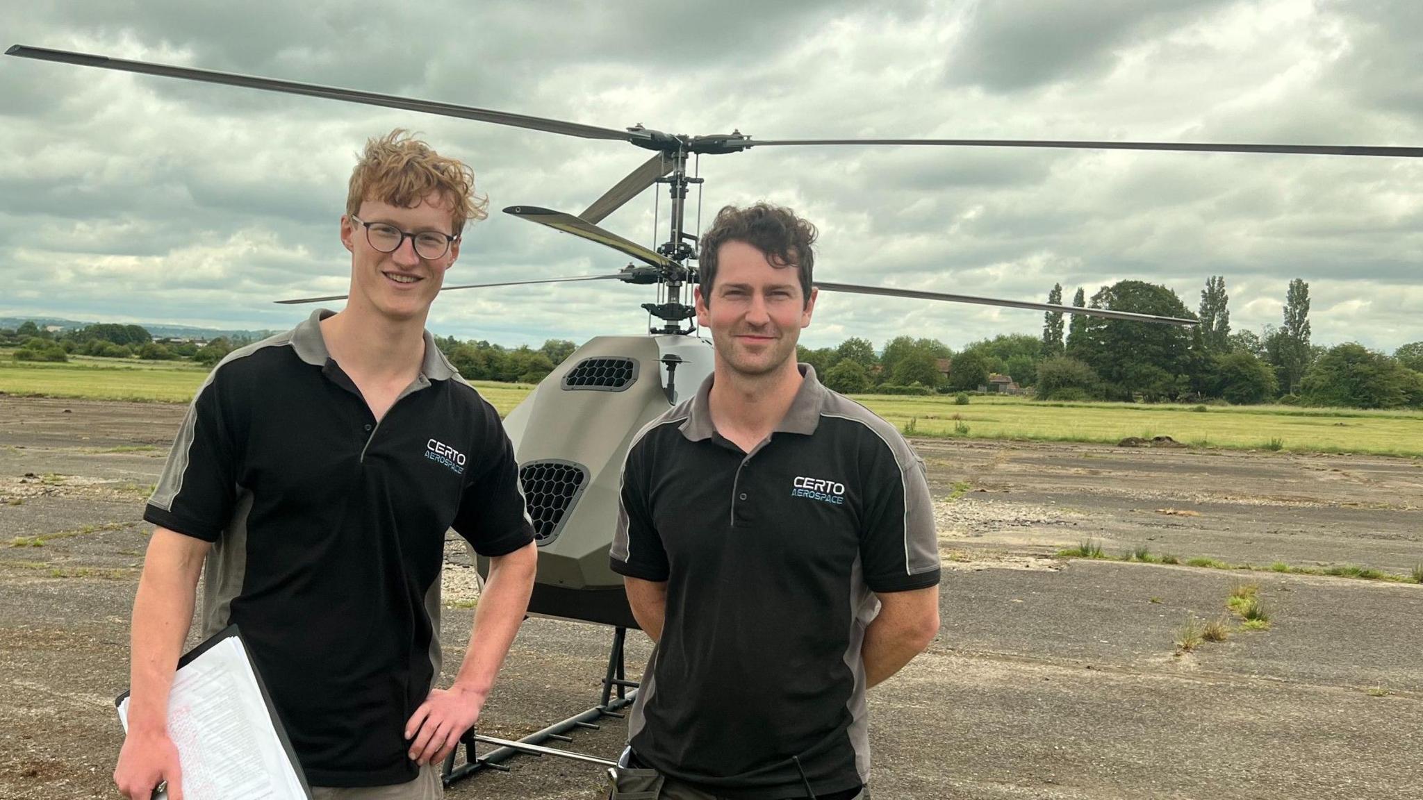 Two young men who are engineers on an unmanned helicopter stand in front of it on Keevil airfield.