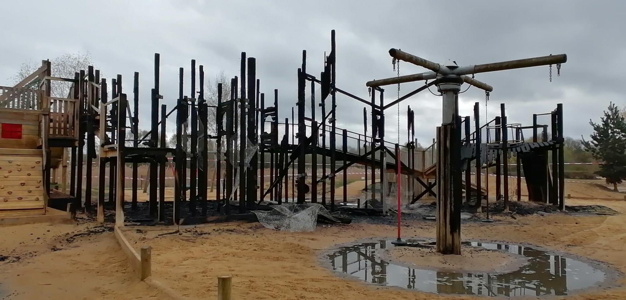 A large play area that has been burnt and left badly damaged. It shows water on the ground, and a wooden structure that has partially collapsed.