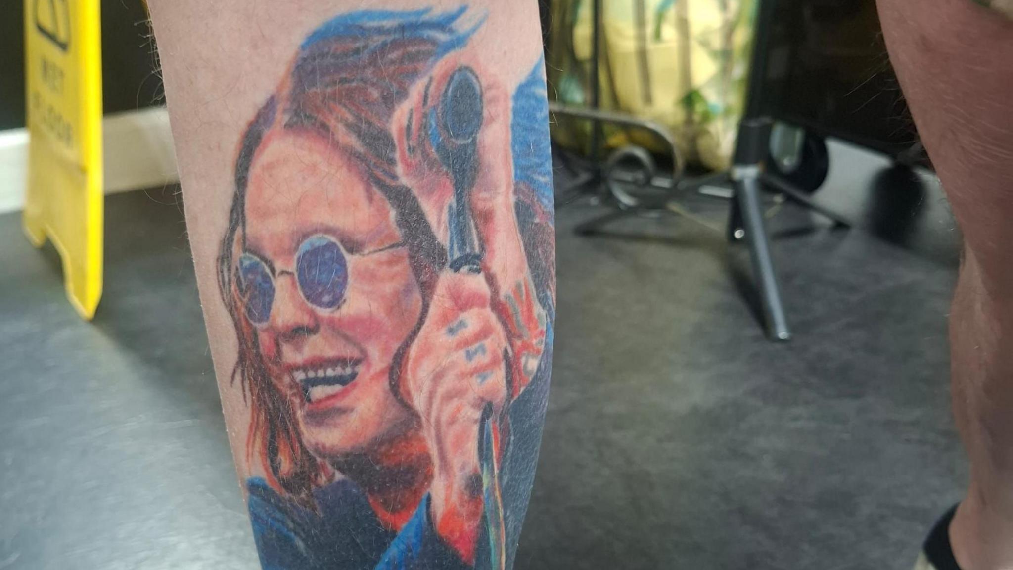 A tattoo of a smiling Ozzy Osbourne. In the image he is waring blue round glasses and a blue jacket and red top. Ozzy is also holding a microphone and has a tattoo on his fingers. 