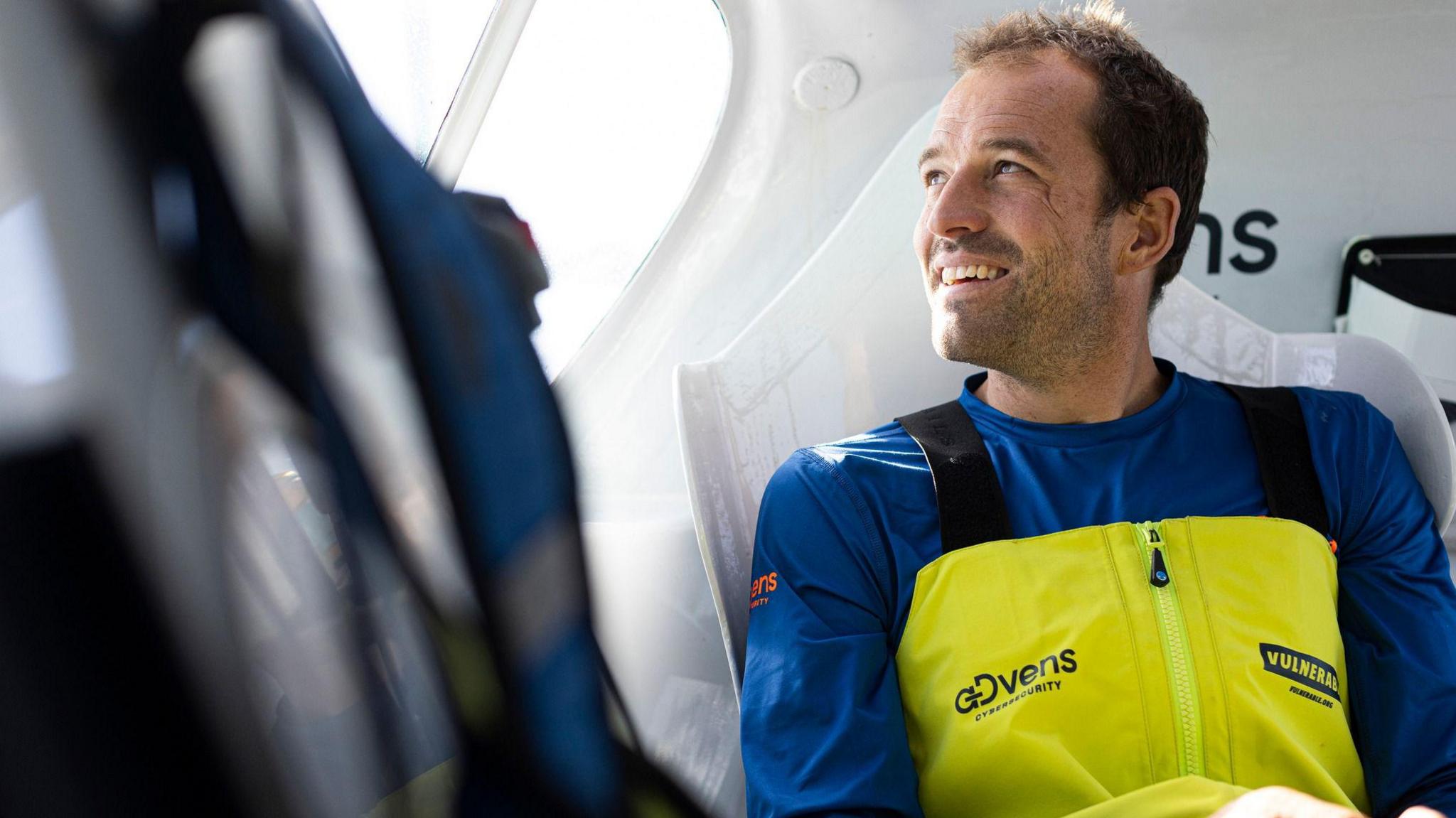 A bearded man in a blue and yellow sailing outfit in the cockpit of a yacht.