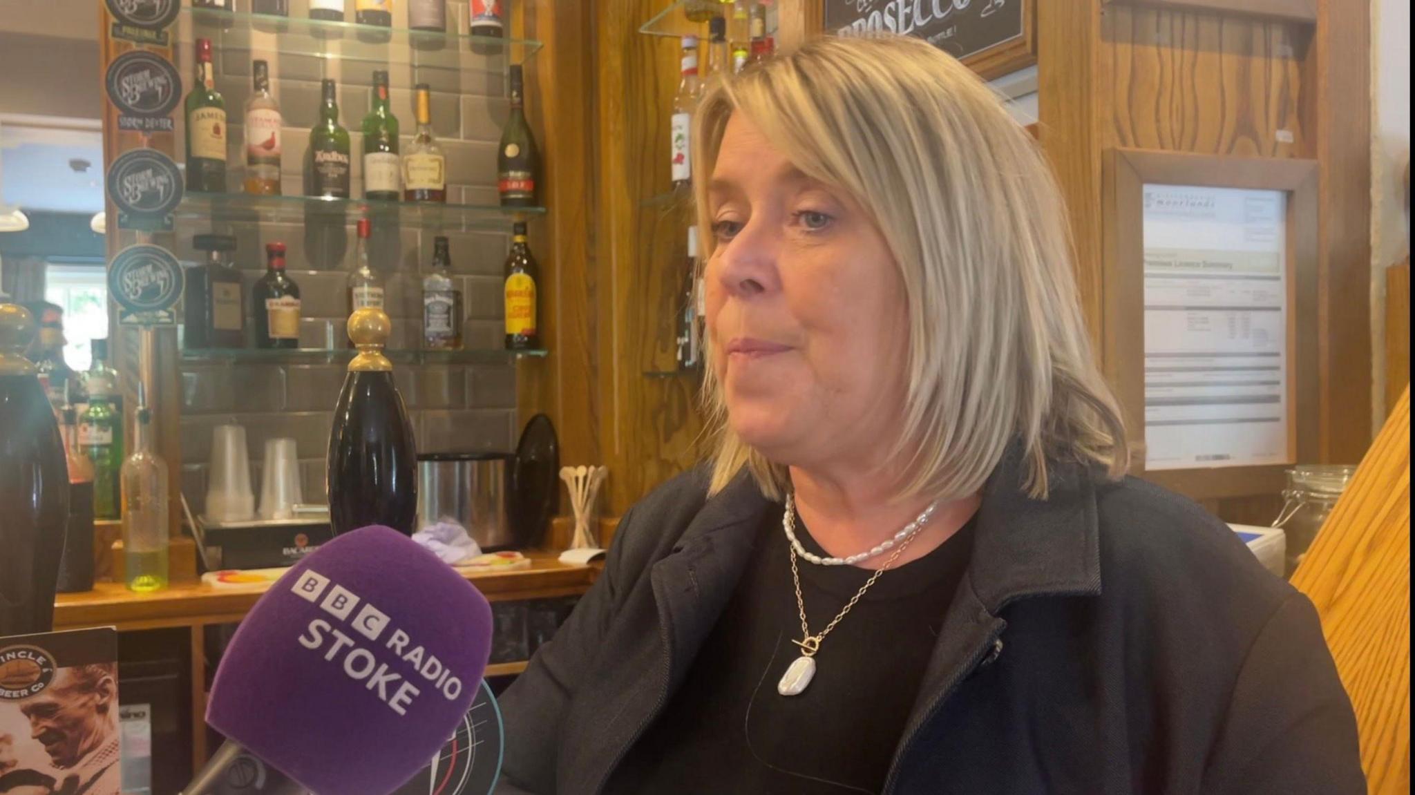 A woman in a black jacket stood in front of a bar with bottles in the background