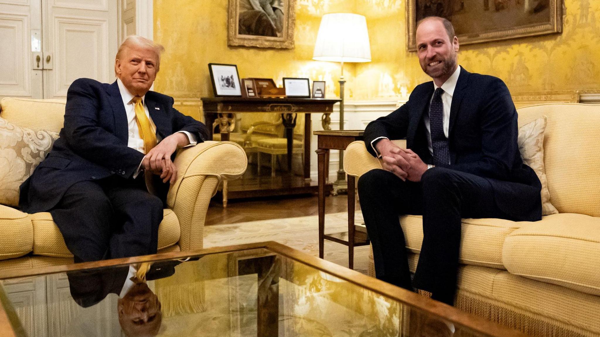 Donald Trump and Prince William seated on cream-colored sofas in a formal living room at the UK ambassador’s residence in Paris, with a glass coffee table in the foreground, ornate yellow wallpaper, and framed photographs on a table in the background.