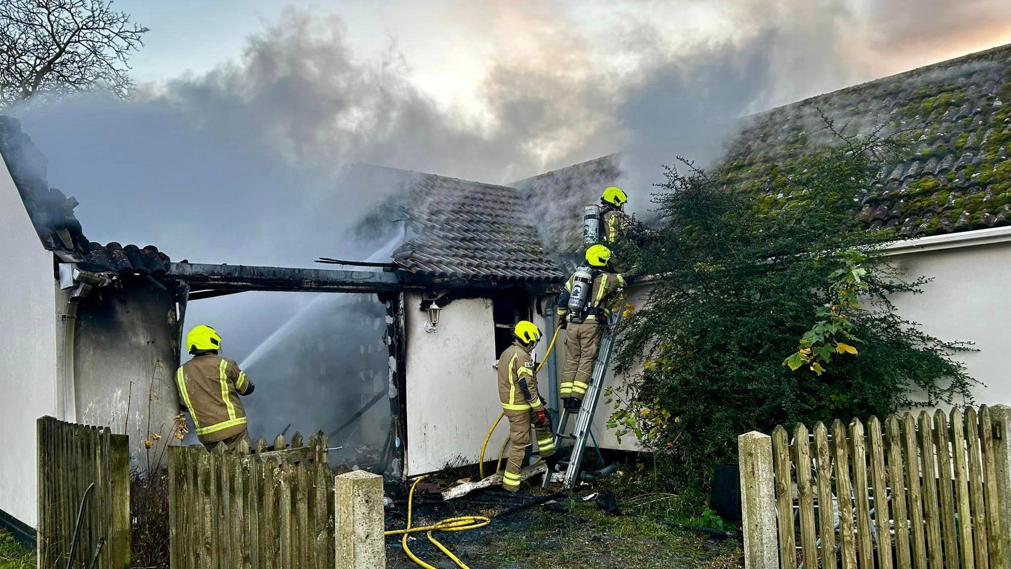 Firefighters are pictured tackling a fire at a property. Two can be seen standing on a small ladder while the tackle the flames and another can be seen holding the ladder steady. A fourth firefighter works nearby with a hose reel jet aiming the water onto the property's roof.