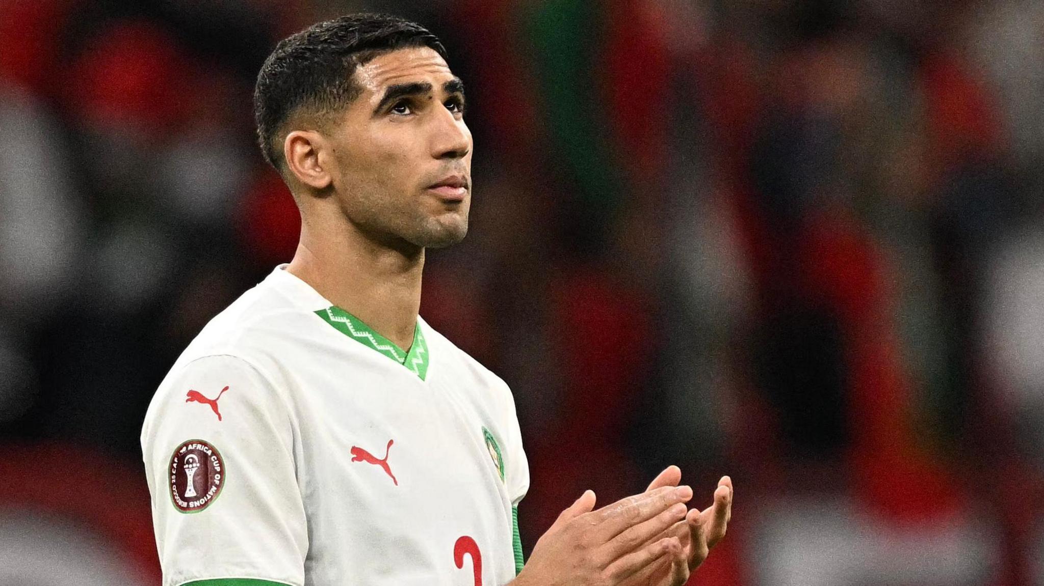 Achraf Hakimi, wearing a white Morocco shirt with green trim, is seen applauding while gazing his eyes skywards