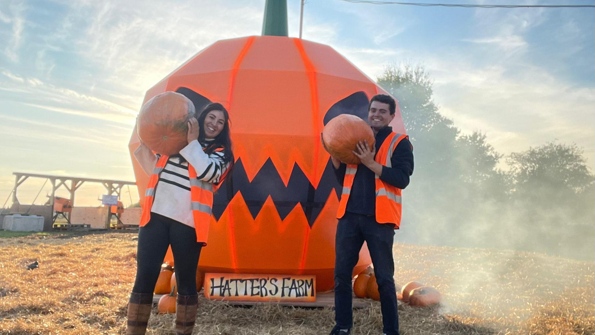 A woman and a man holding a pumpkin each and smile at the camera. Behind them is a smokey blown up pumpkin which is one of the attractions at the farm. They are photographed outside in a field and they are both wearing high-visibility jackets. 