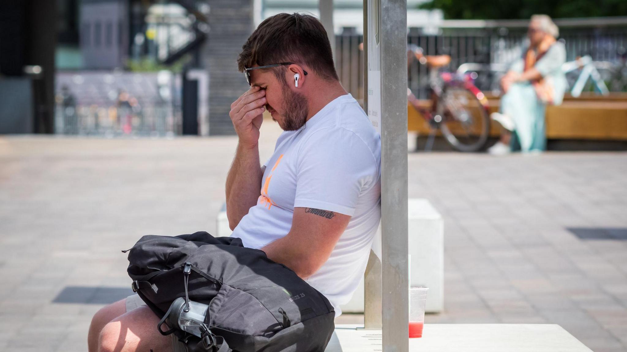 A man in a white t shirt is sitting next to a black rucksack and rubbing his eyes. He looks exhausted by the heat.