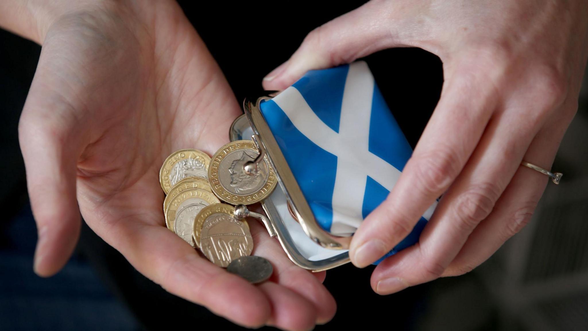 Coins being emptied out of a purse with a Saltire on it into someone's hands.