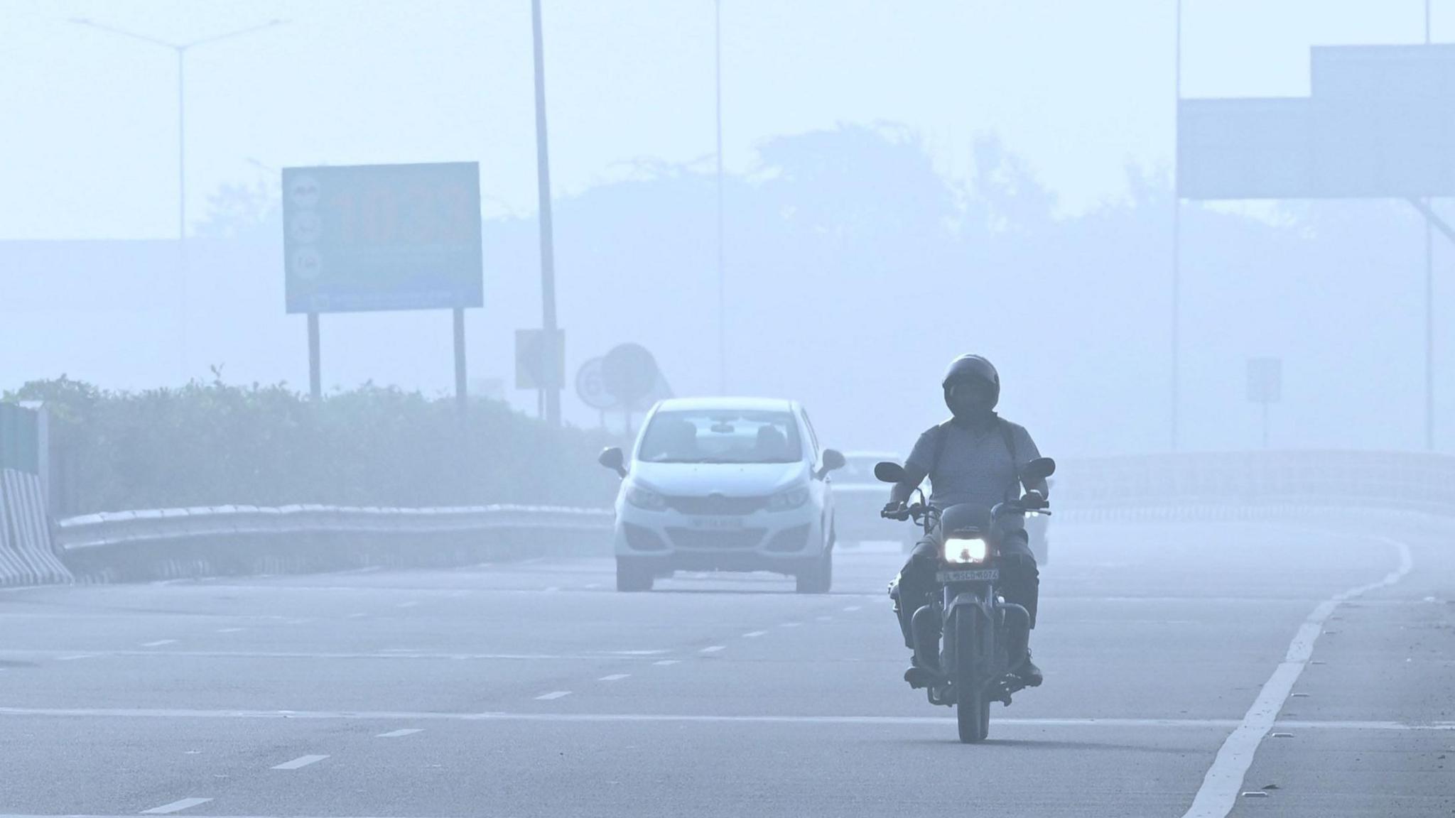 A man riding on a bike on a road in India's capital. The air is hazy suggesting smog.