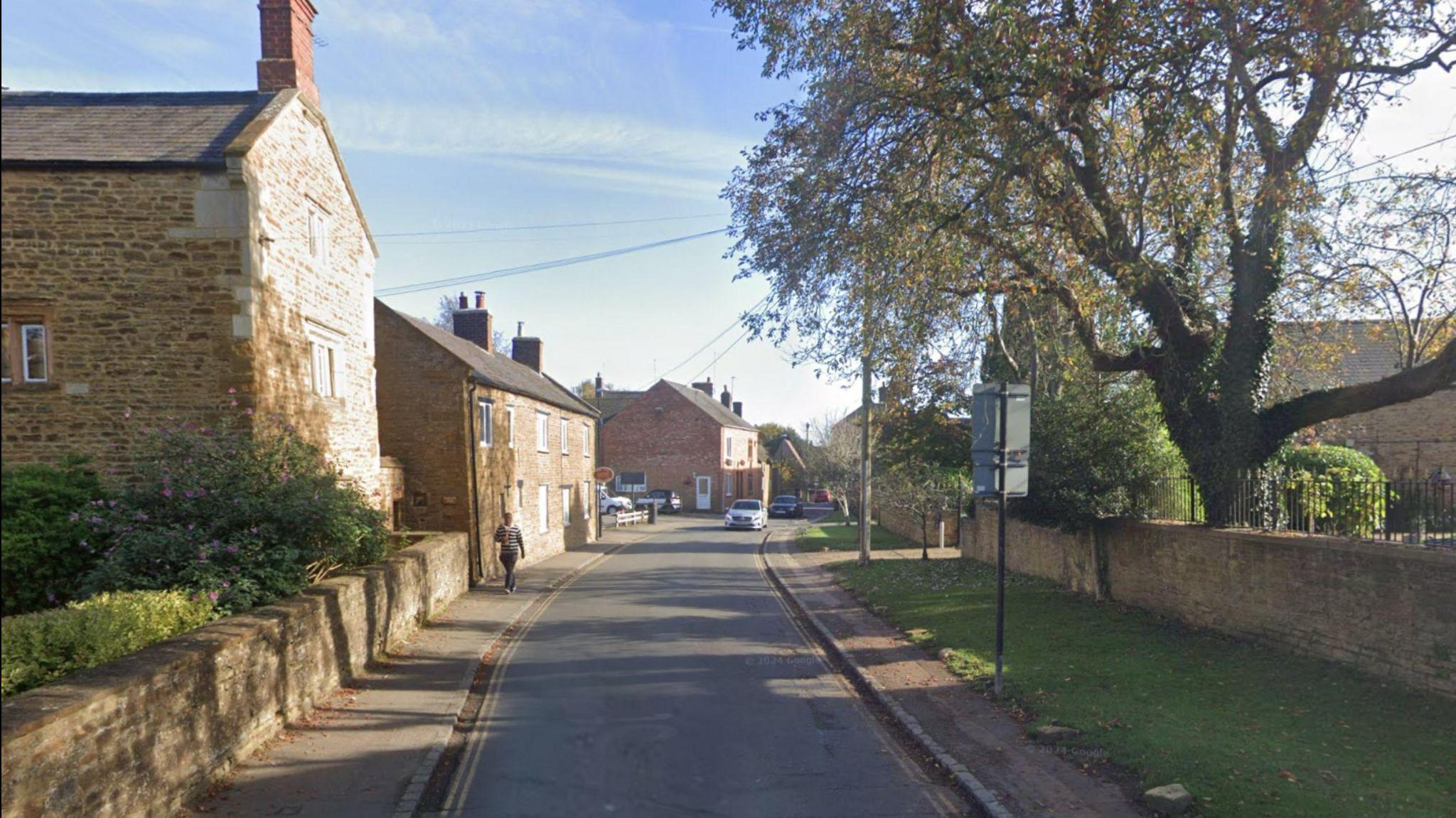 A road through a rural village. There is a pavement either side, beyond which is a grass verge on one side and a stone wall on the other.  Two-storey stone houses are visible on the left.  A person is walking down the pavement in the distance.