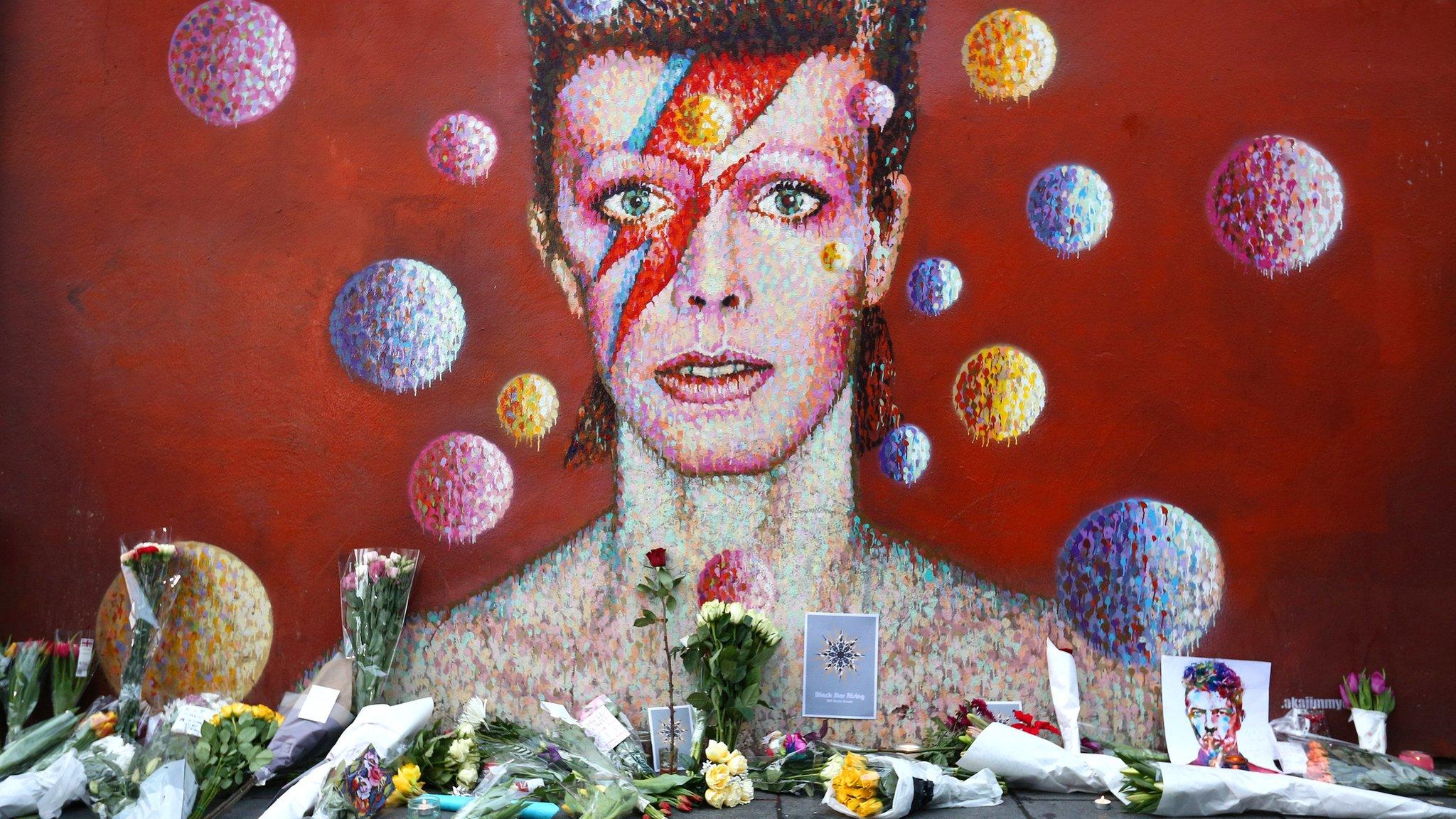 Flowers are laid in front of a mural of David Bowie