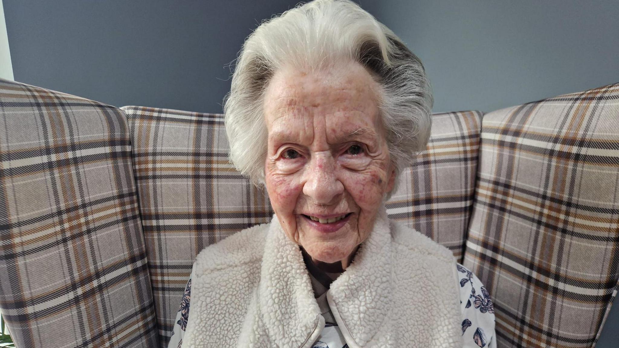 An elderly woman, 104-year-old, Mary Smith smiles into the camera. She has white hair, with dark streaks and is wearing a cosy cream gilet over a patterned top.
