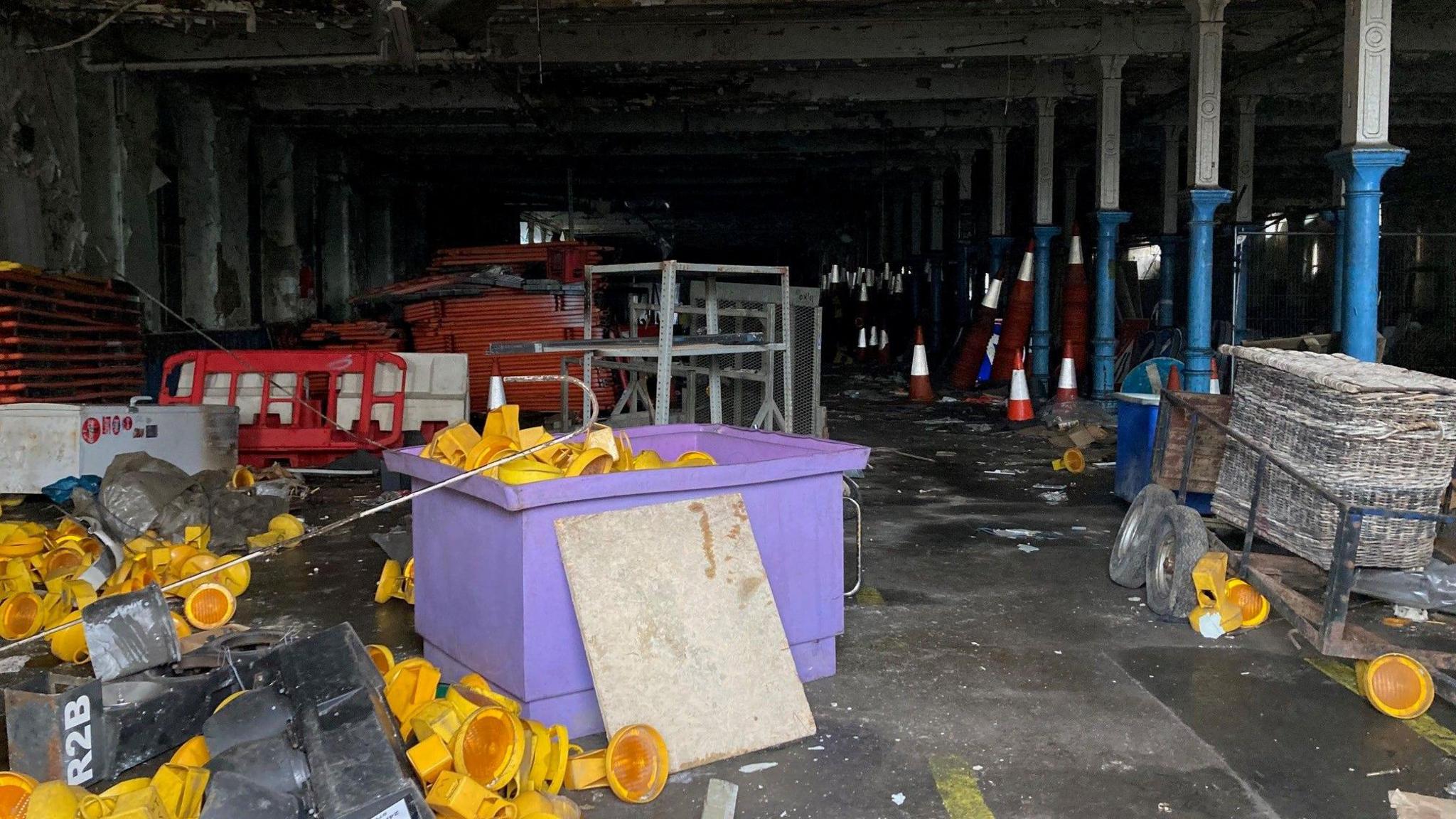 Yellow roadwork lights piled on the floor and in a purple plastic container inside a large, downstairs room inside the mill. Red and white traffic cones and and safety barriers can be seen in the background.  
 